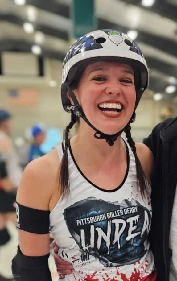 A woman wearing a helmet and athletic attire smiling at a roller derby event, with the shirt reading "Pittsburgh Roller Derby Unk".