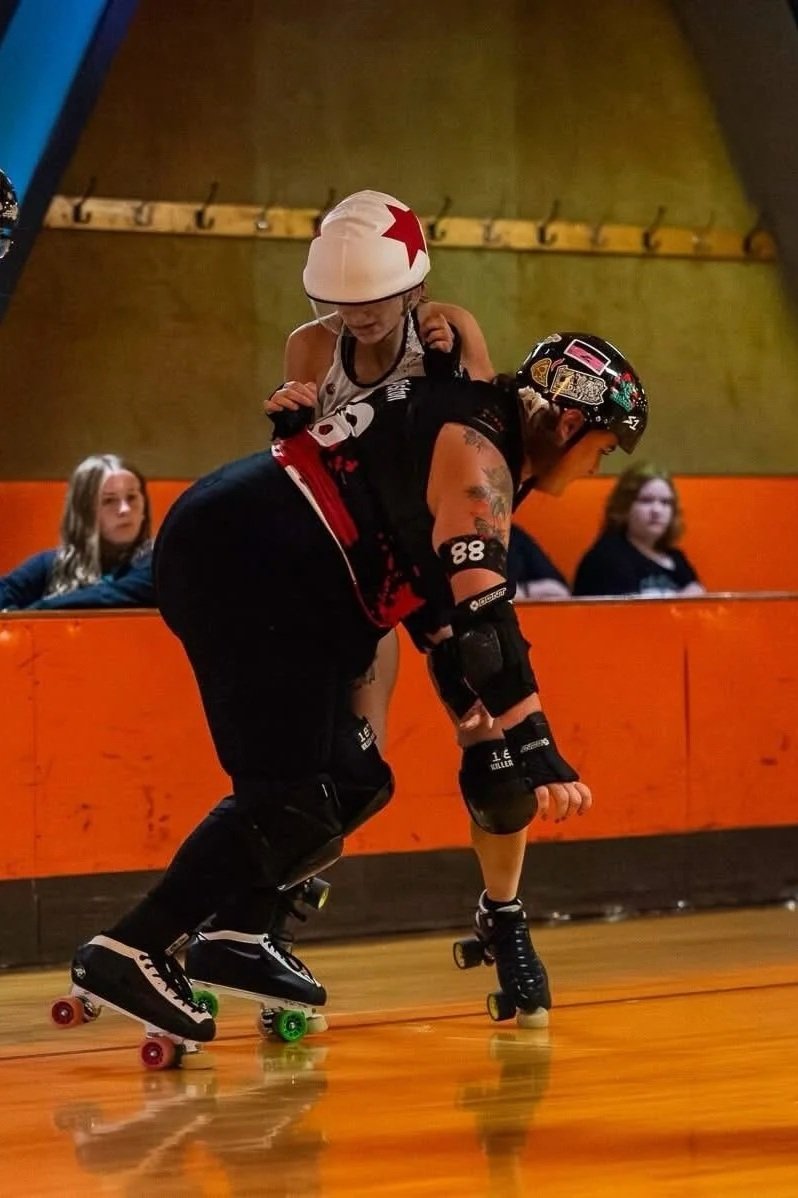 A person wearing a helmet and roller derby gear bends down on roller skates, with a child riding on their back, in an indoor roller rink.