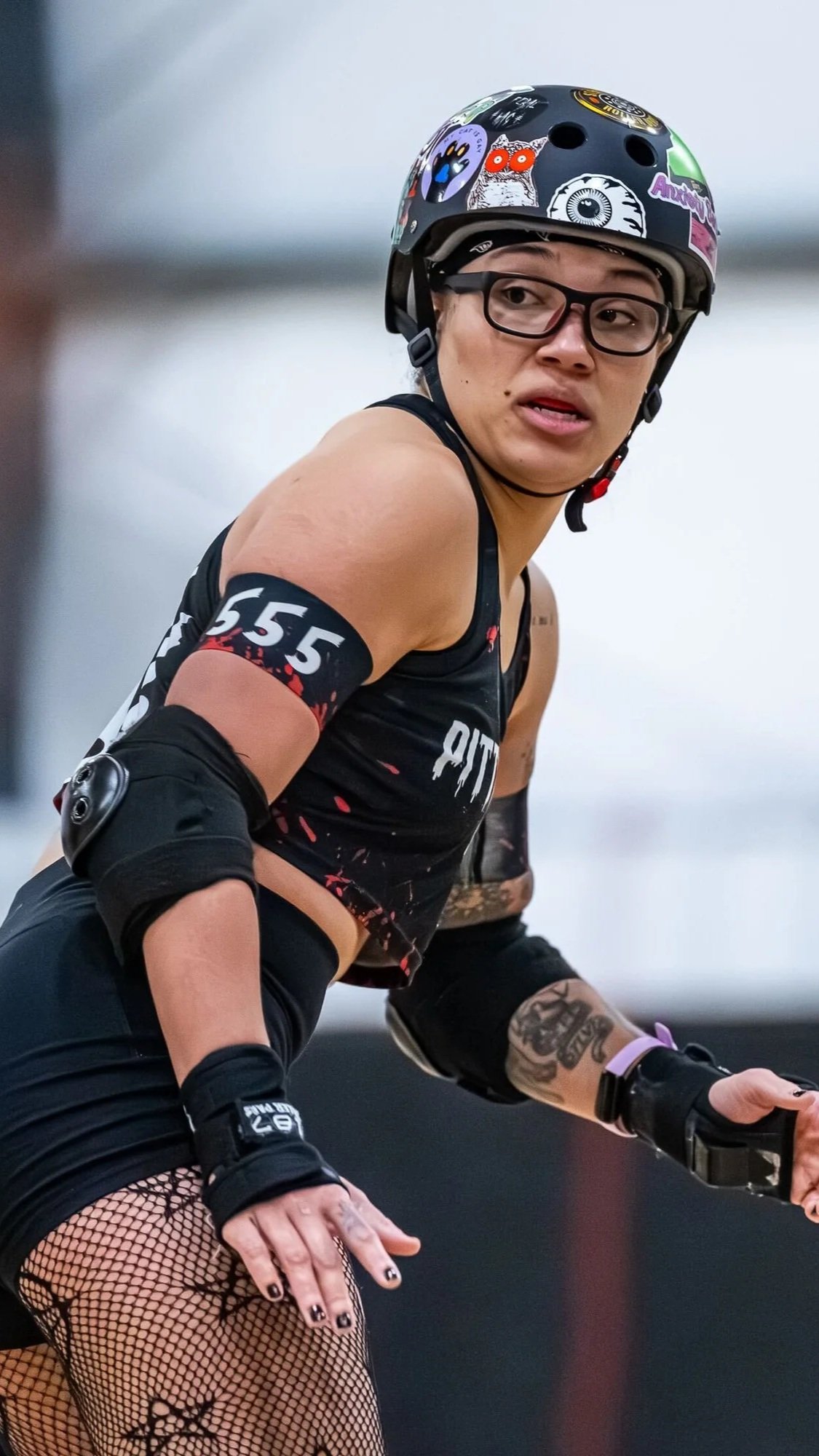 A female inline skater wearing a black helmet with stickers, glasses, a black sleeveless top with red accents, fishnet stockings, and protective gear, skating indoors.