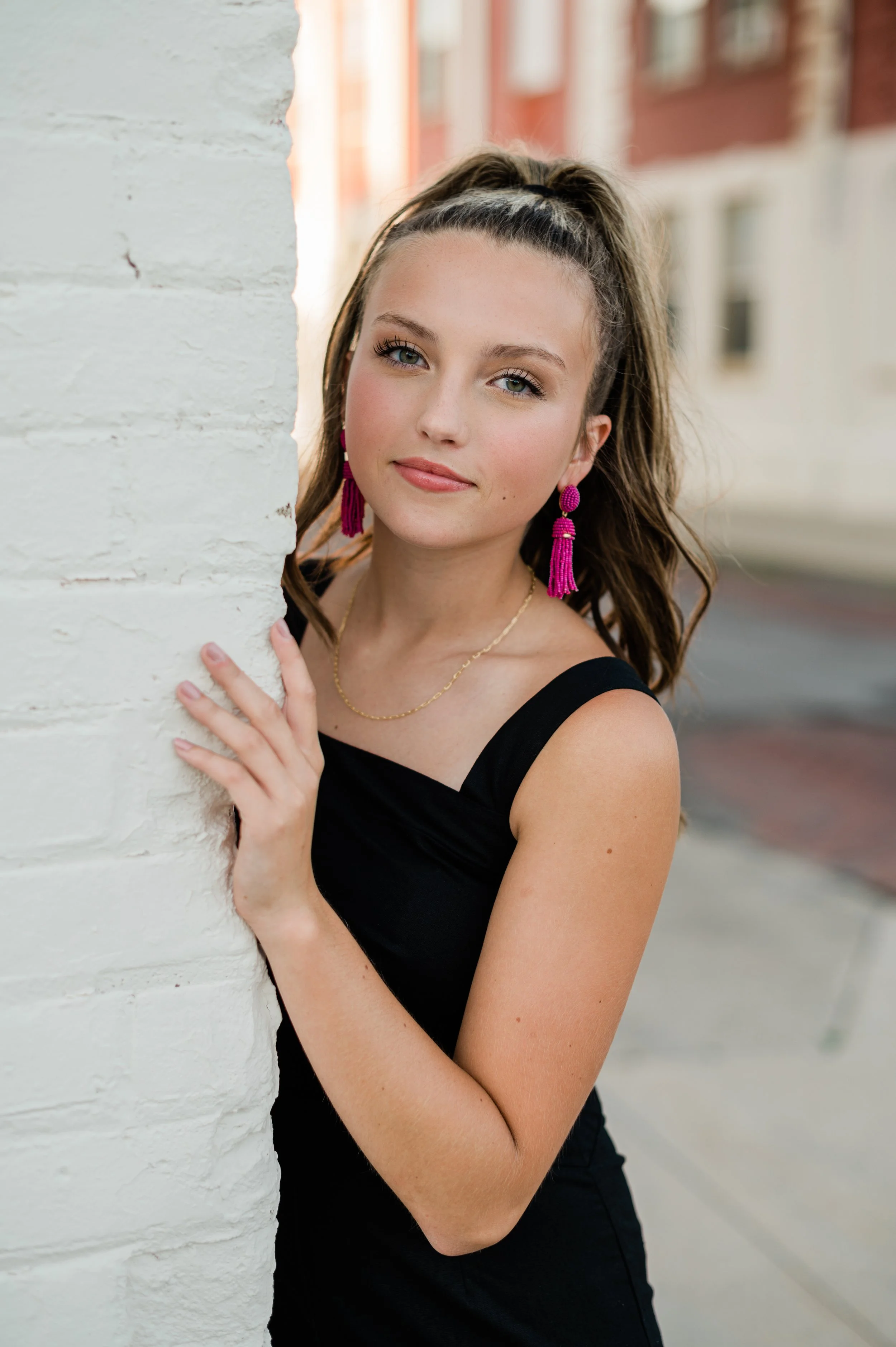 Young woman with long brown hair and blue eyes peeking from behind a white brick wall, wearing a black sleeveless top and pink earrings, outdoors with blurred buildings in the background.