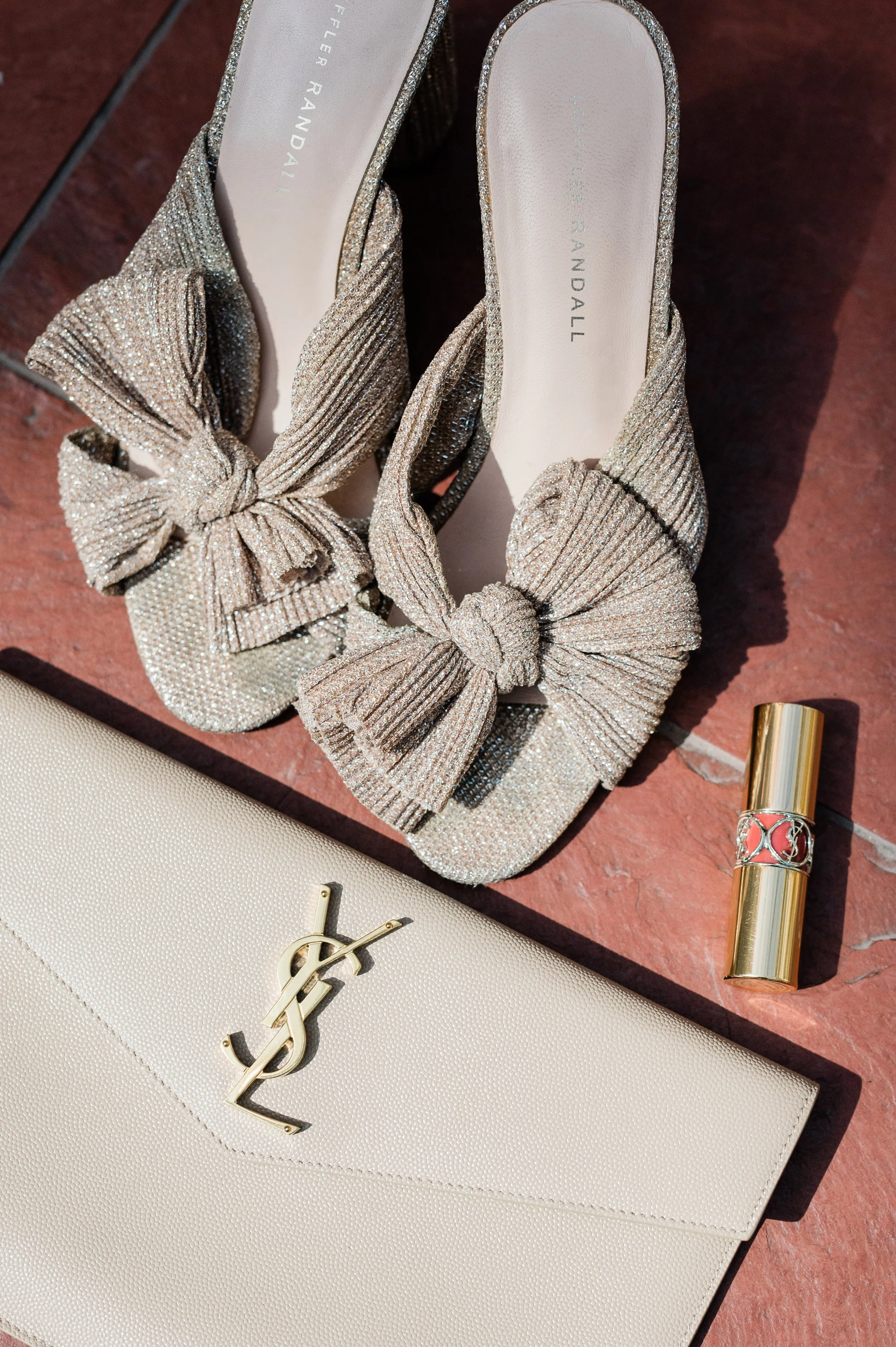 A pair of glittering beige high heels with large bow embellishments, a beige Yves Saint Laurent clutch with gold logo, and a gold lipstick tube with a decorative band, all arranged on a reddish surface.