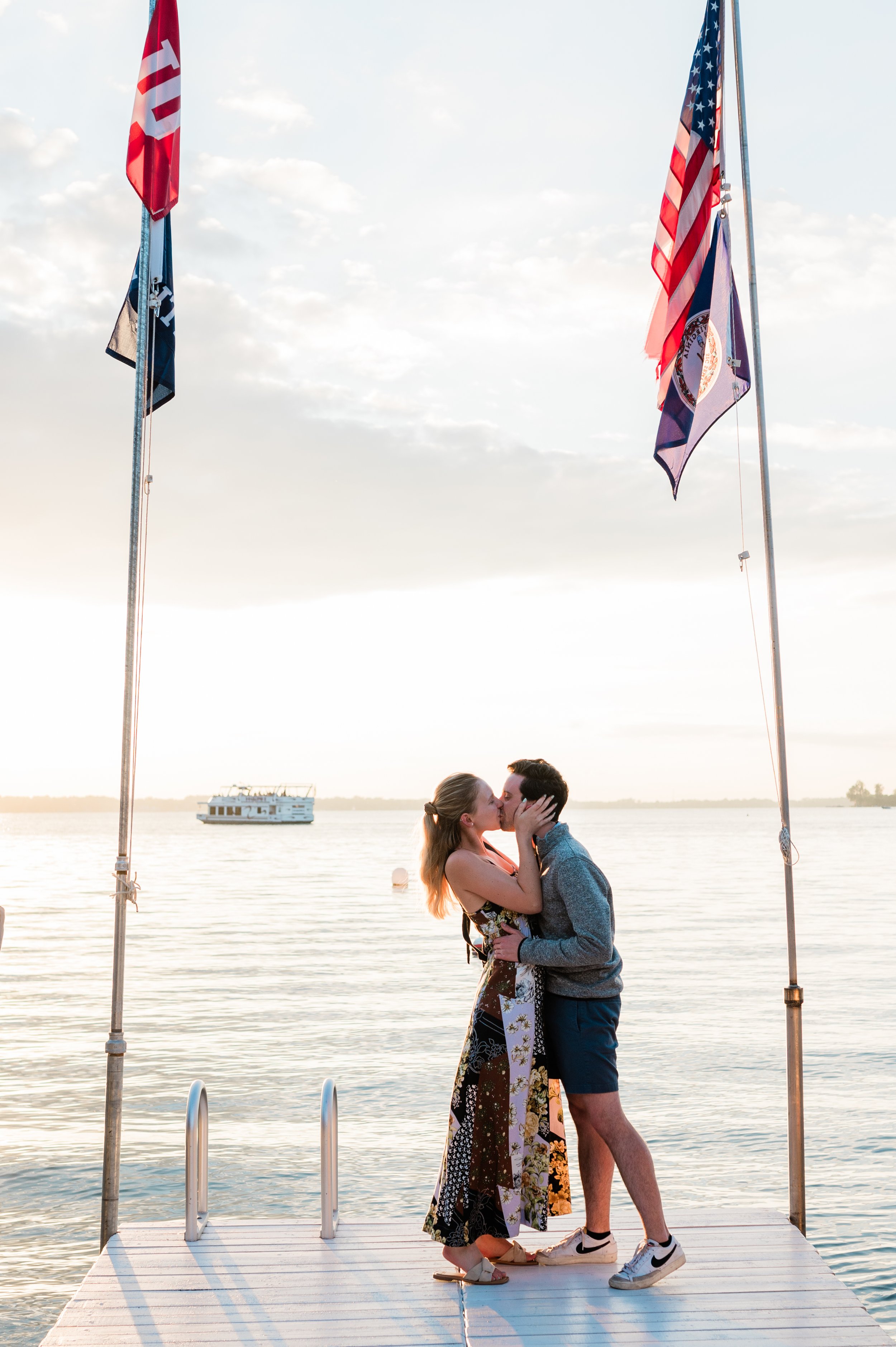 A couple kissing on a dock at sunset, with the water and a boat in the background, and American flags on either side of them.