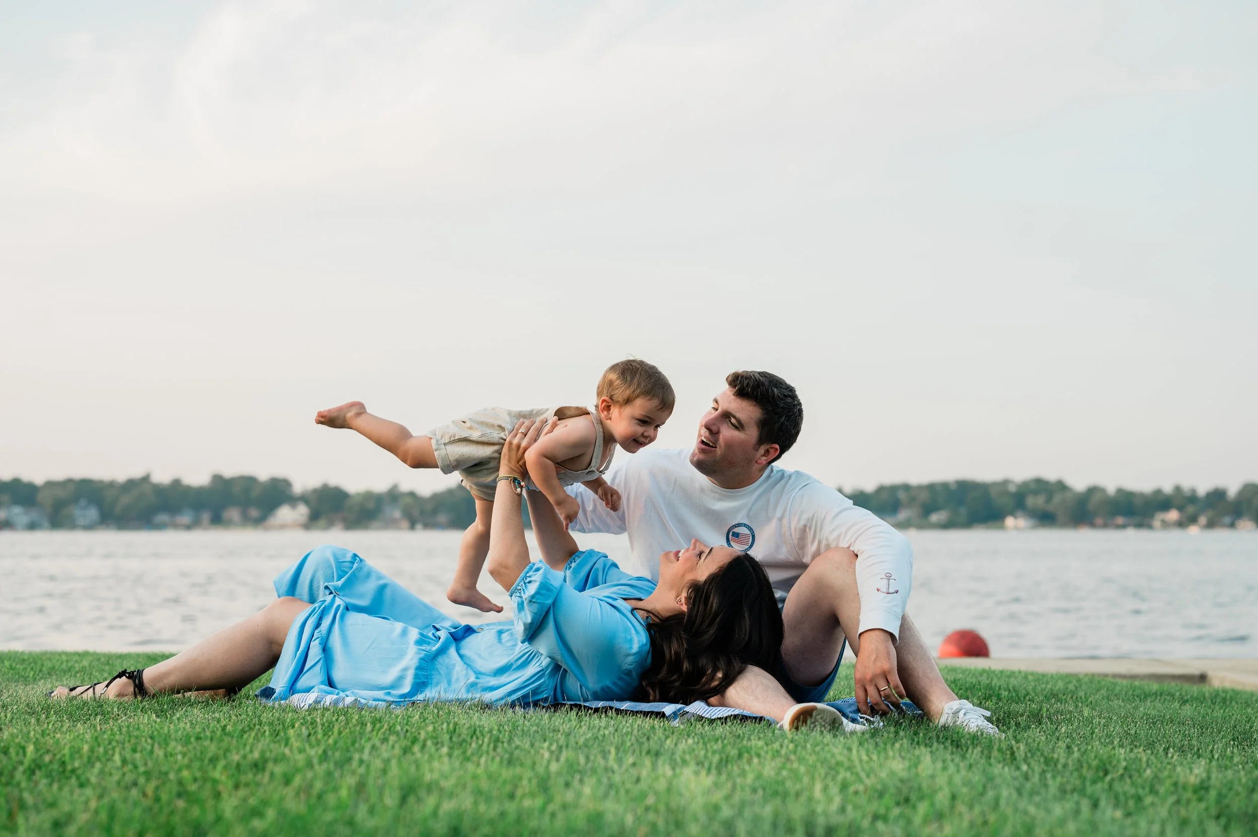 A family of three enjoying a day by the water, with the father sitting on the grass, the mother lying down, and the young son being lifted in the air, all smiling and interacting happily.
