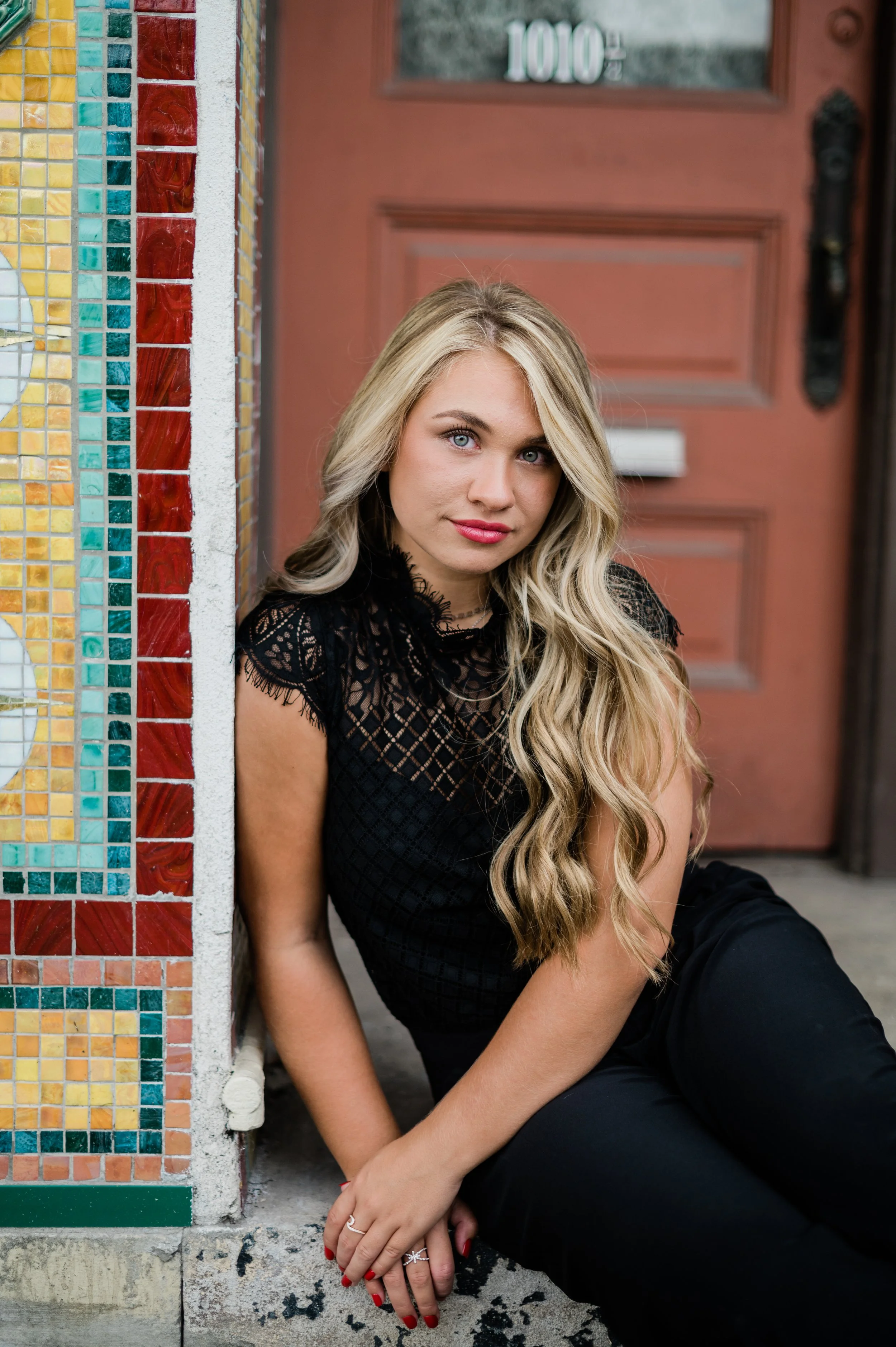 Young woman with long, wavy blonde hair and light blue eyes, wearing a black lace top, sitting on the ground near a colorful mosaic wall and a red door in Fort Wayne, Indiana.
