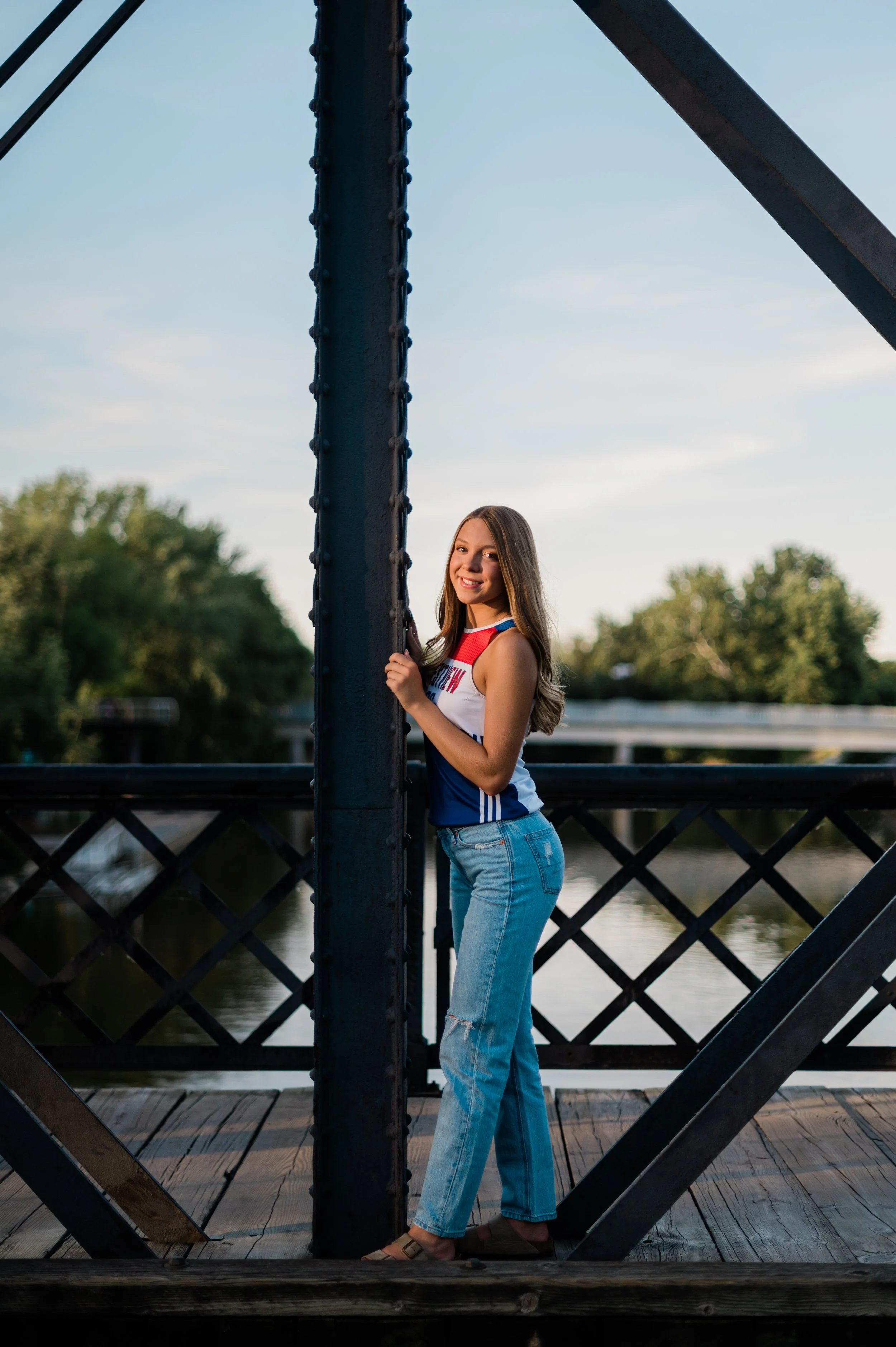A smiling young woman with long hair stands on a wooden bridge near a body of water, holding onto a vertical metal post on the Wells Street Bridge in Fort Wayne, Indiana.