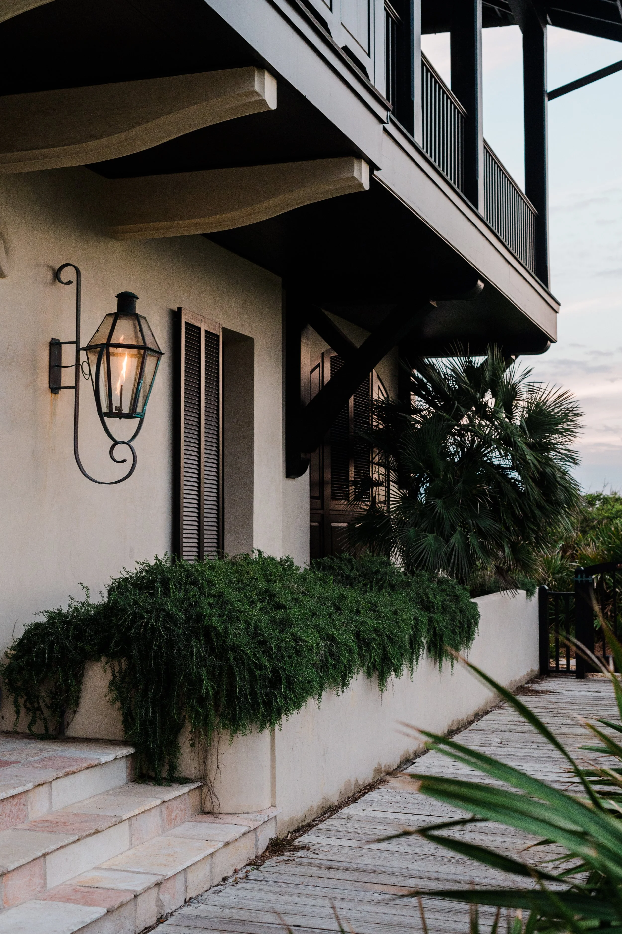 Exterior of a house with a wall-mounted lantern, dark shutters, lush greenery, and a wooden pathway at dusk.