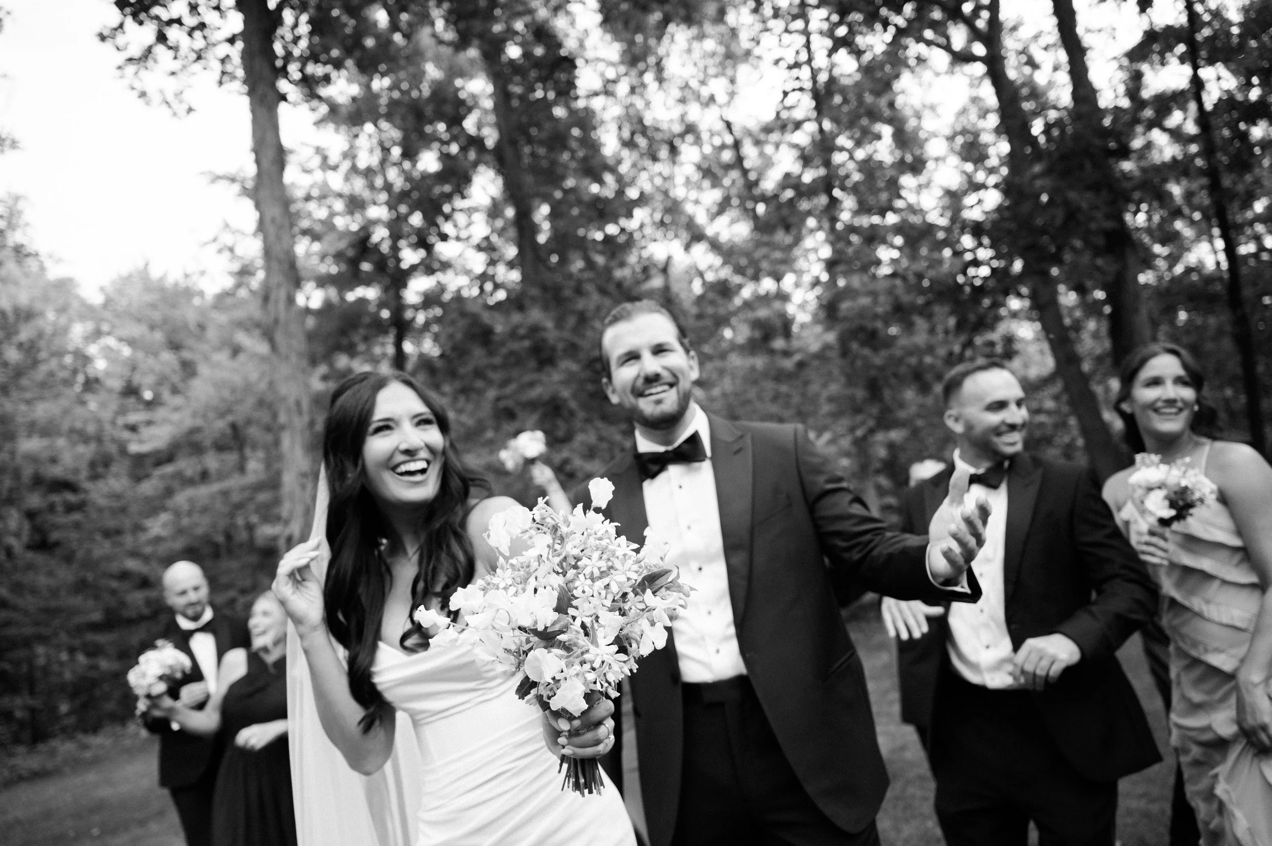 Bride and groom dancing joyfully toward the camera during their wedding at The Bluffs at Conner Prairie in Indianapolis, photographed in black and white from a low angle with their wedding party in the background.