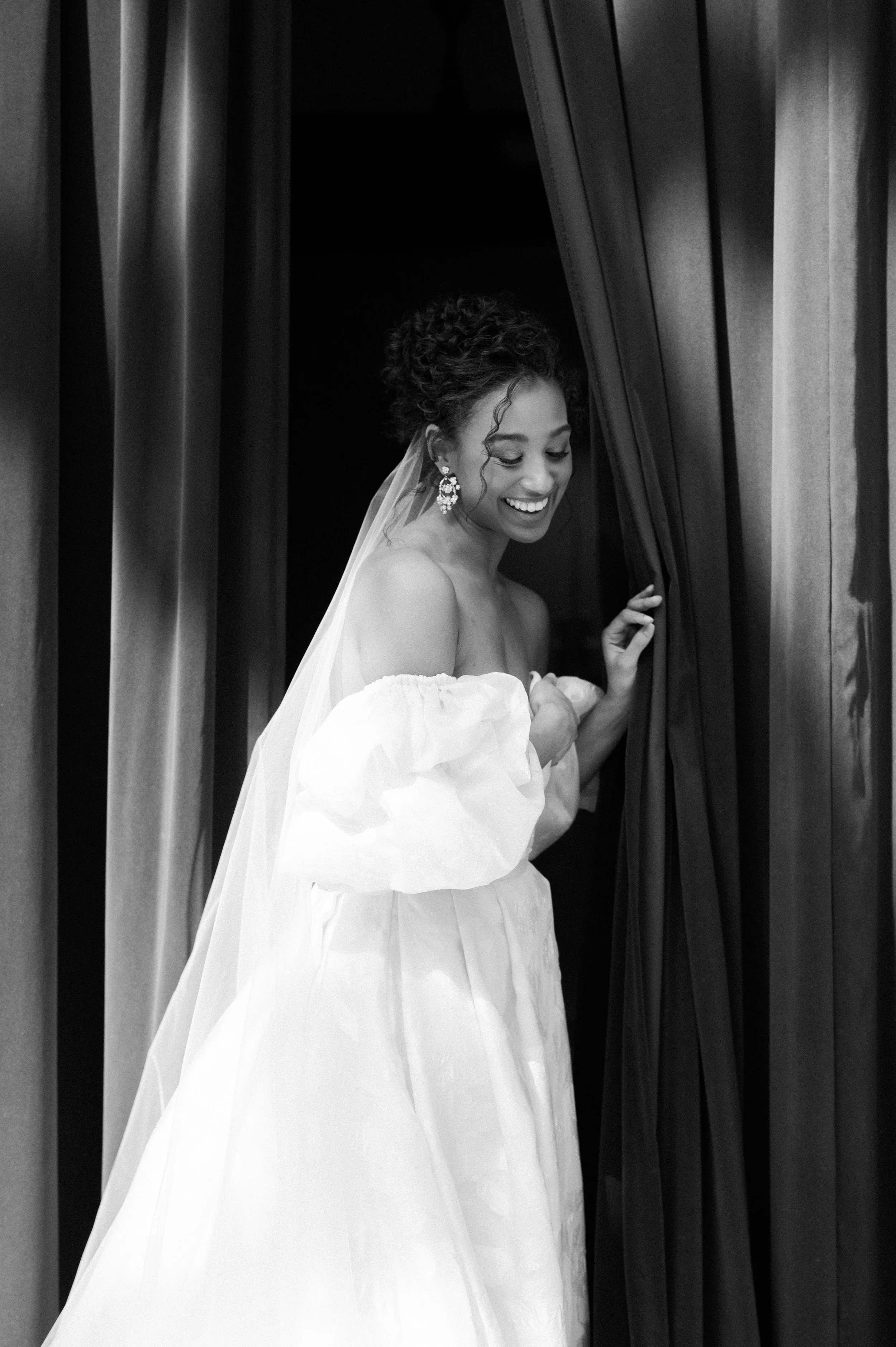 Black and white photo of a smiling woman in a wedding dress, holding the curtain and looking down at the Columbus Museum of Art
