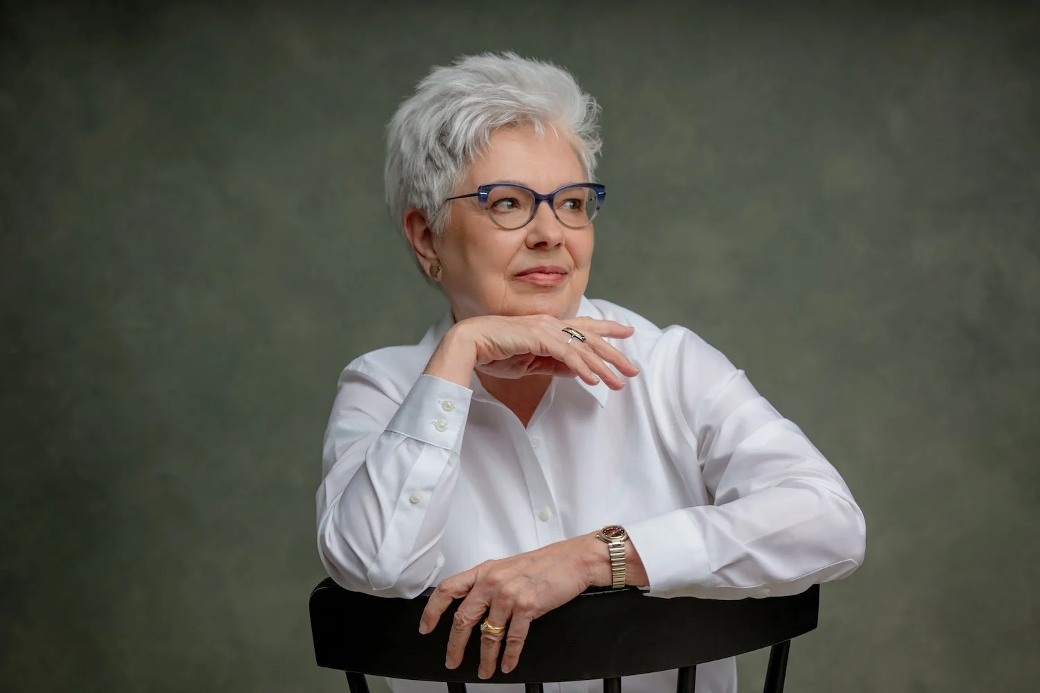 Woman with short silver hair and glasses wearing a white shirt and leaning on a chair during a studio portrait session