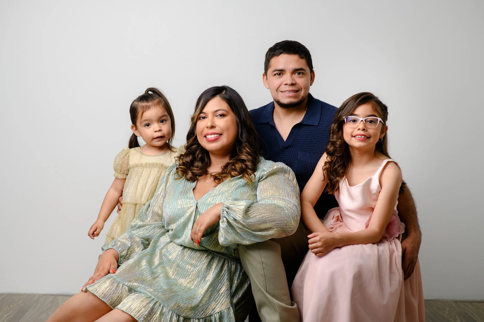 Family of four posing together in a studio, with two young daughters wearing cream and pink dresses