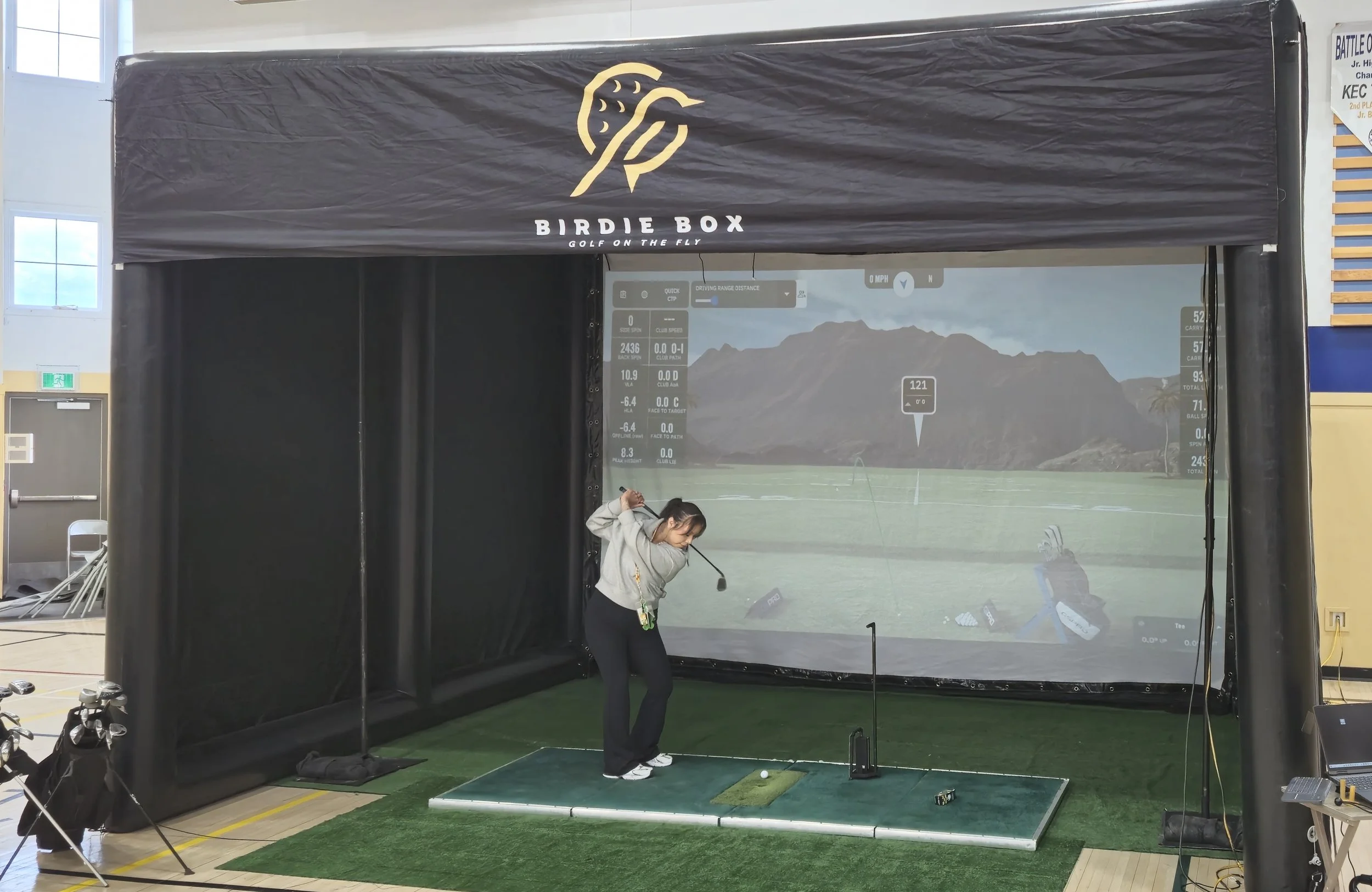 Indoor golf simulator setup with a woman practicing her swing, a large screen displaying a golf course, and golf clubs nearby.