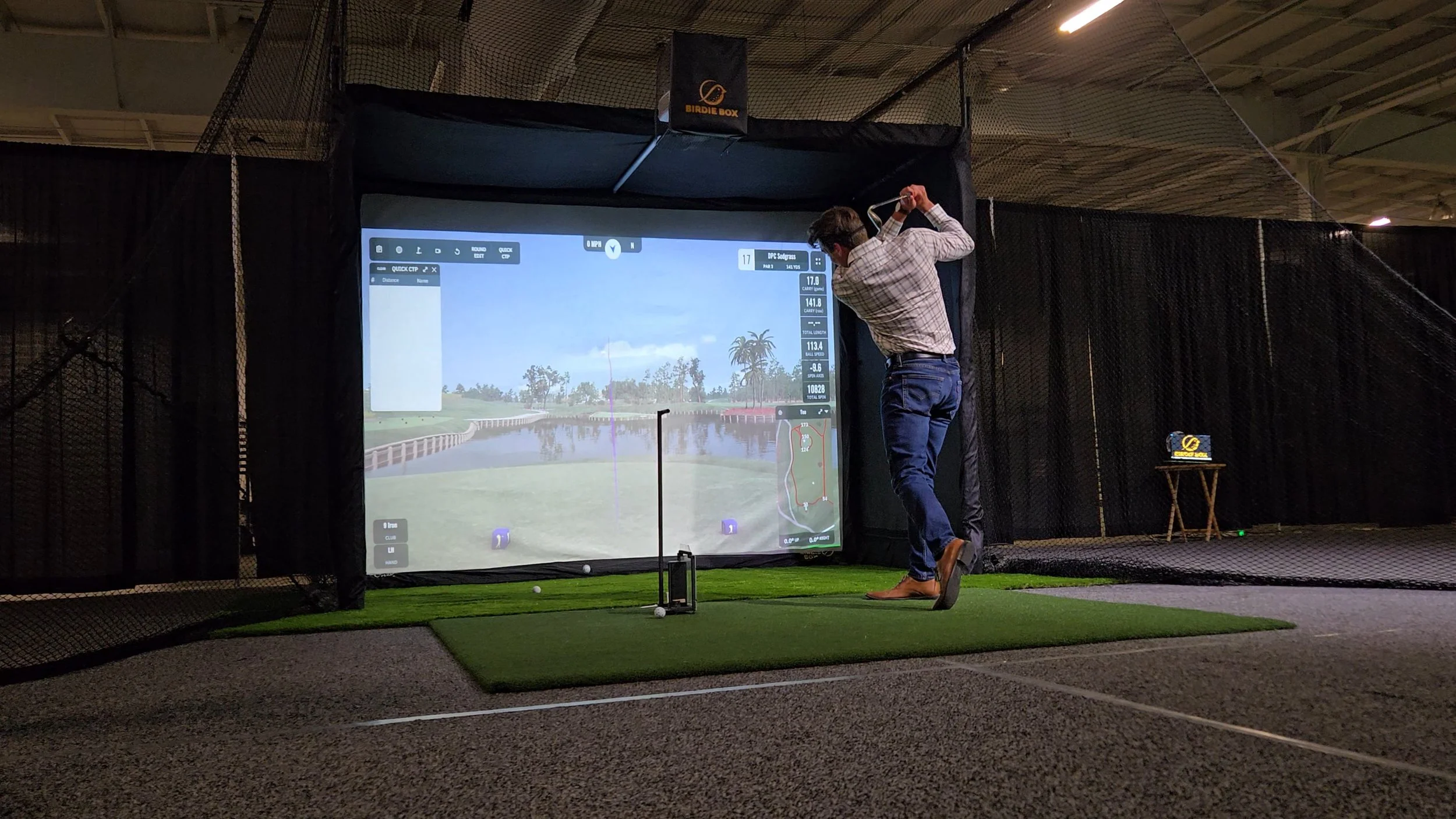 Man in plaid shirt and jeans taking a golf swing in an indoor simulator at a golf center.