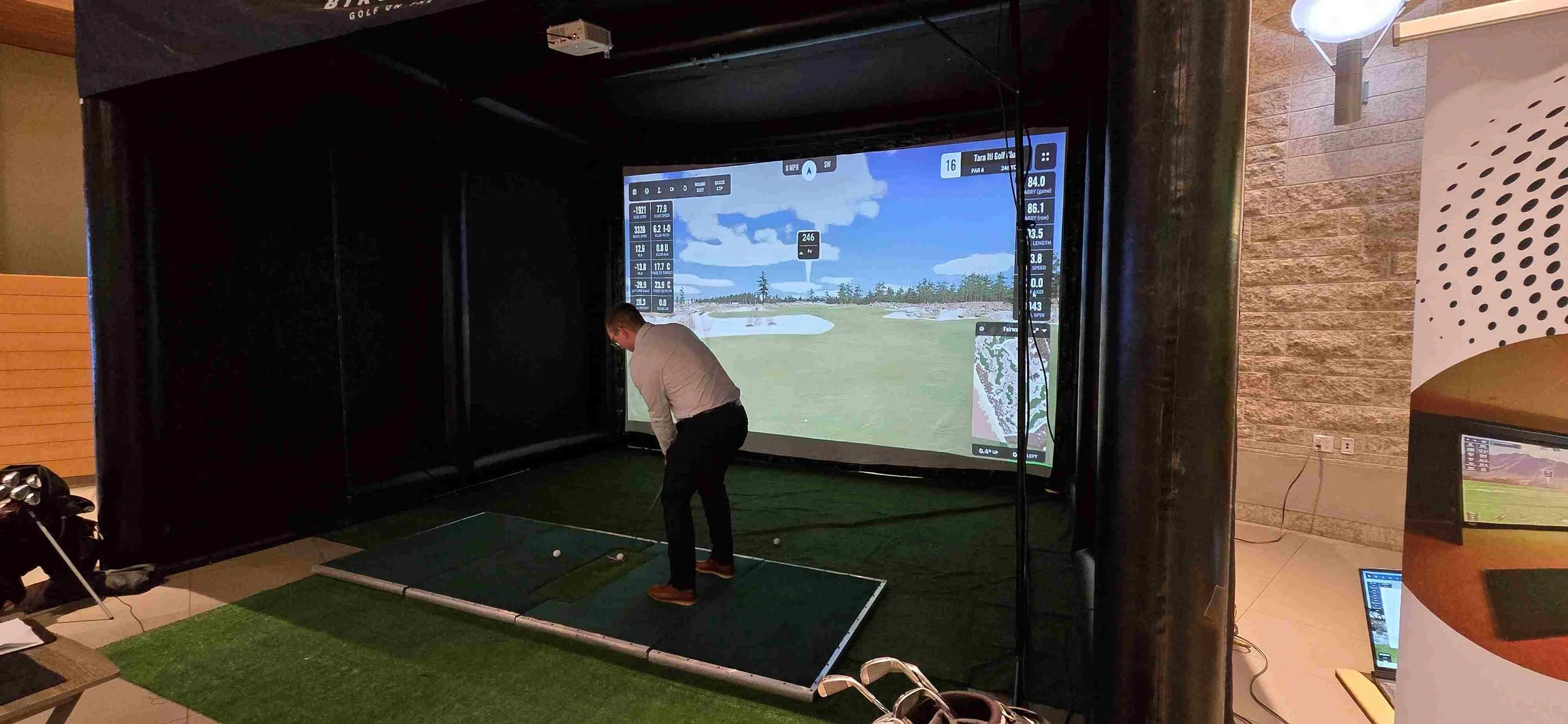 Indoor golf simulation room with a man practicing putting on a small artificial green, facing a large screen displaying a golf course simulation.