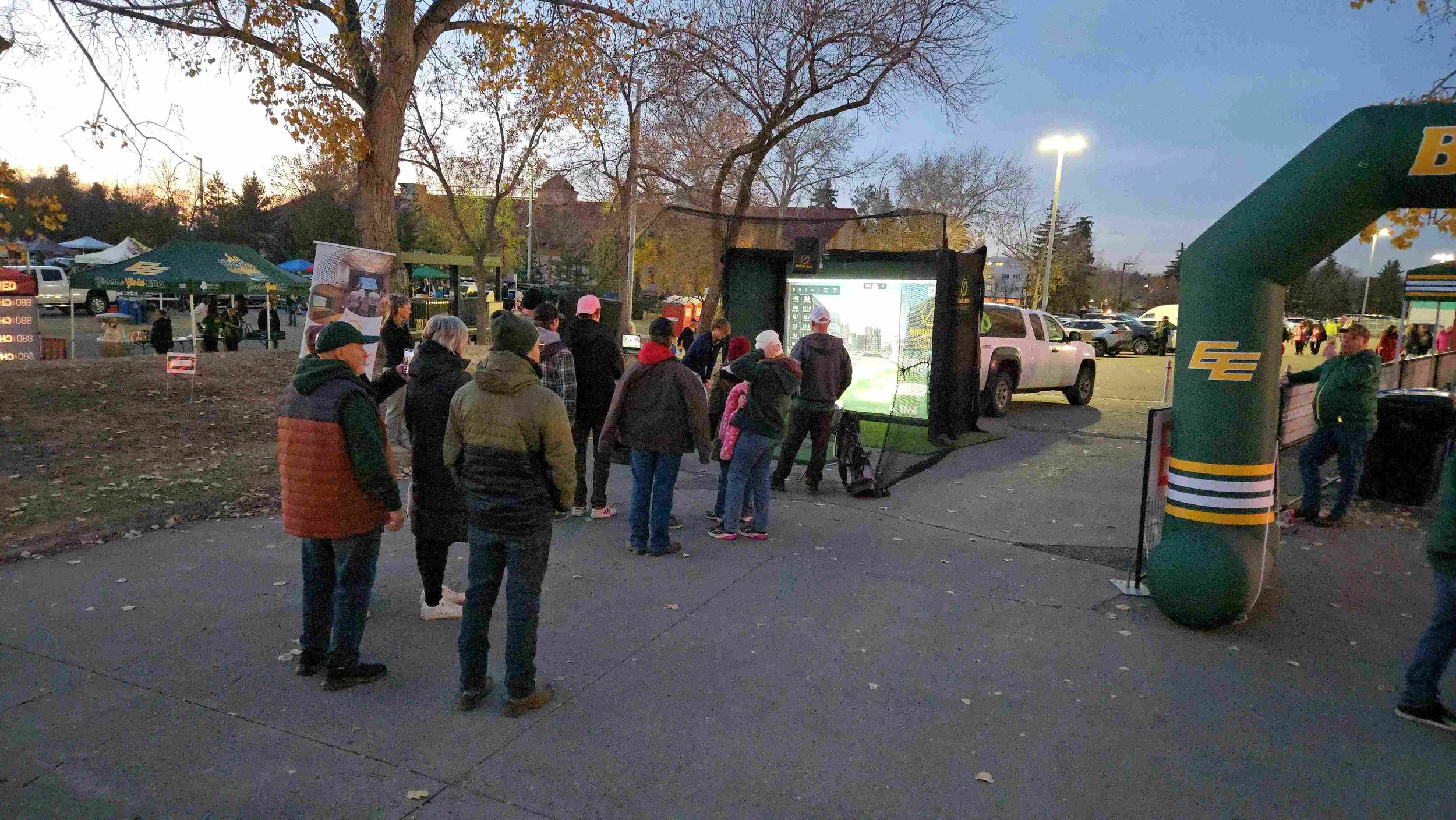 People waiting in line outside to play a golf-themed arcade game at an outdoor event in a parking lot during dusk. There are trees with leafless branches, tents, parked cars, and a large inflatable archway.