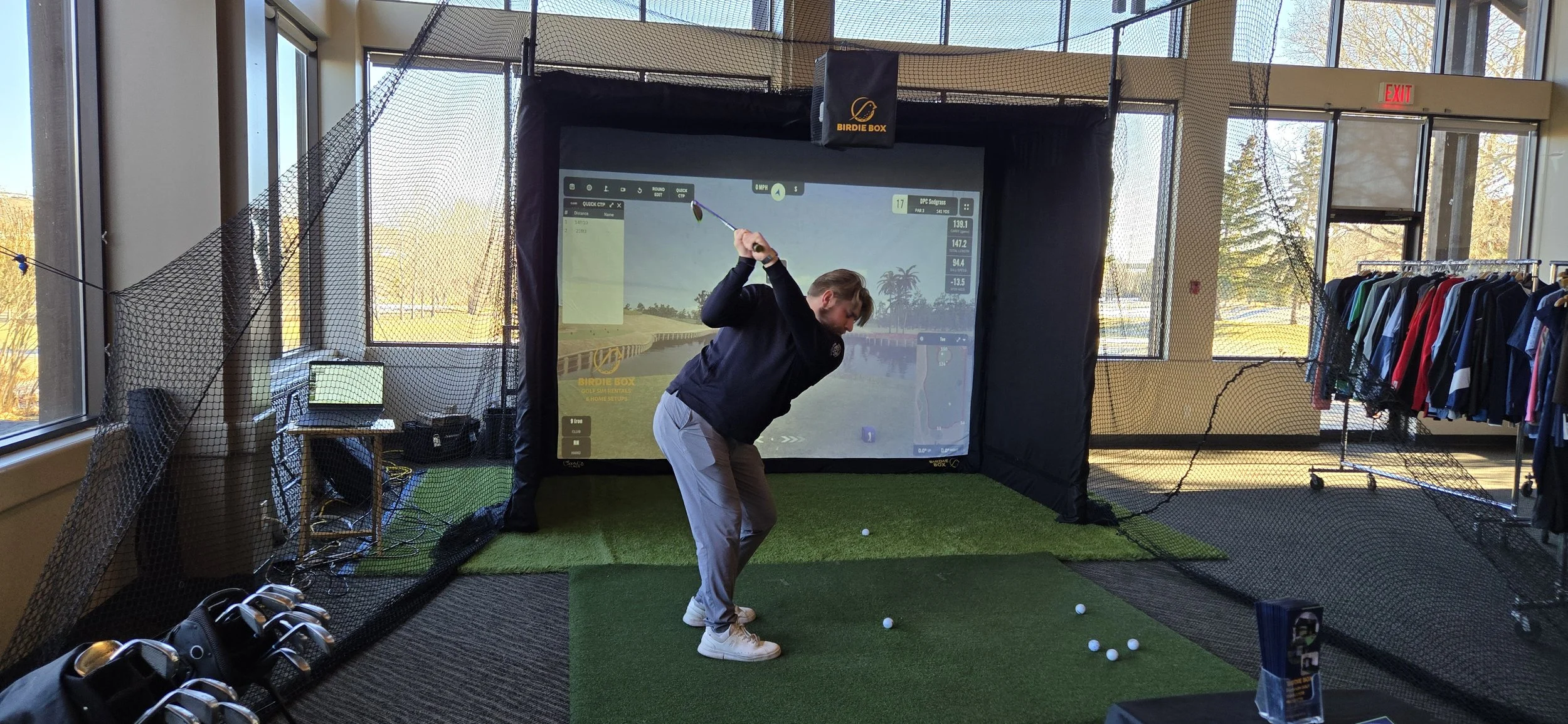 Man practicing golf in indoor simulator with screen showing virtual golf course, golf clubs, and balls on green mat.