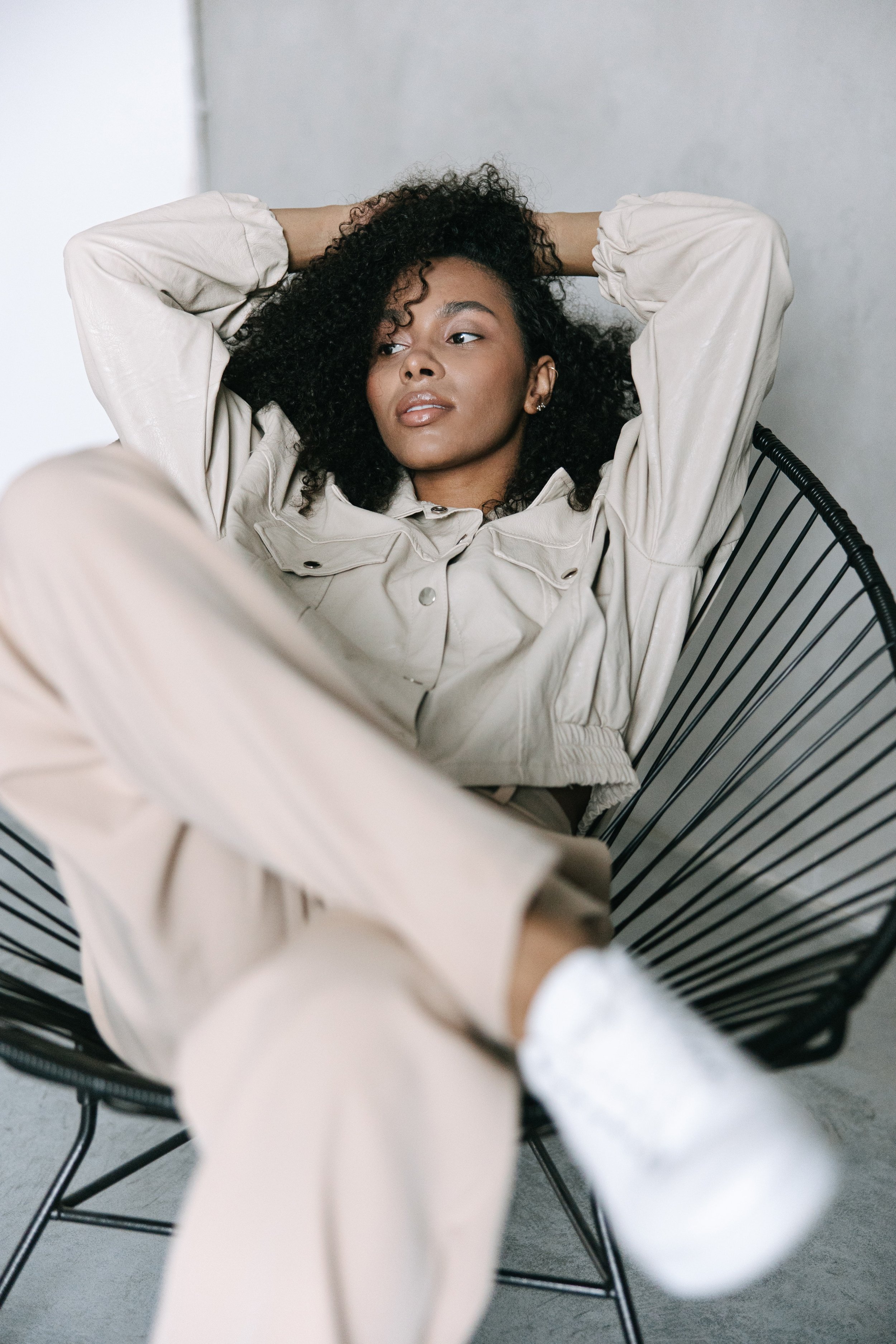A young woman with curly hair relaxing in a black wire chair, wearing a beige outfit, with one arm behind her head and another resting on her lap.