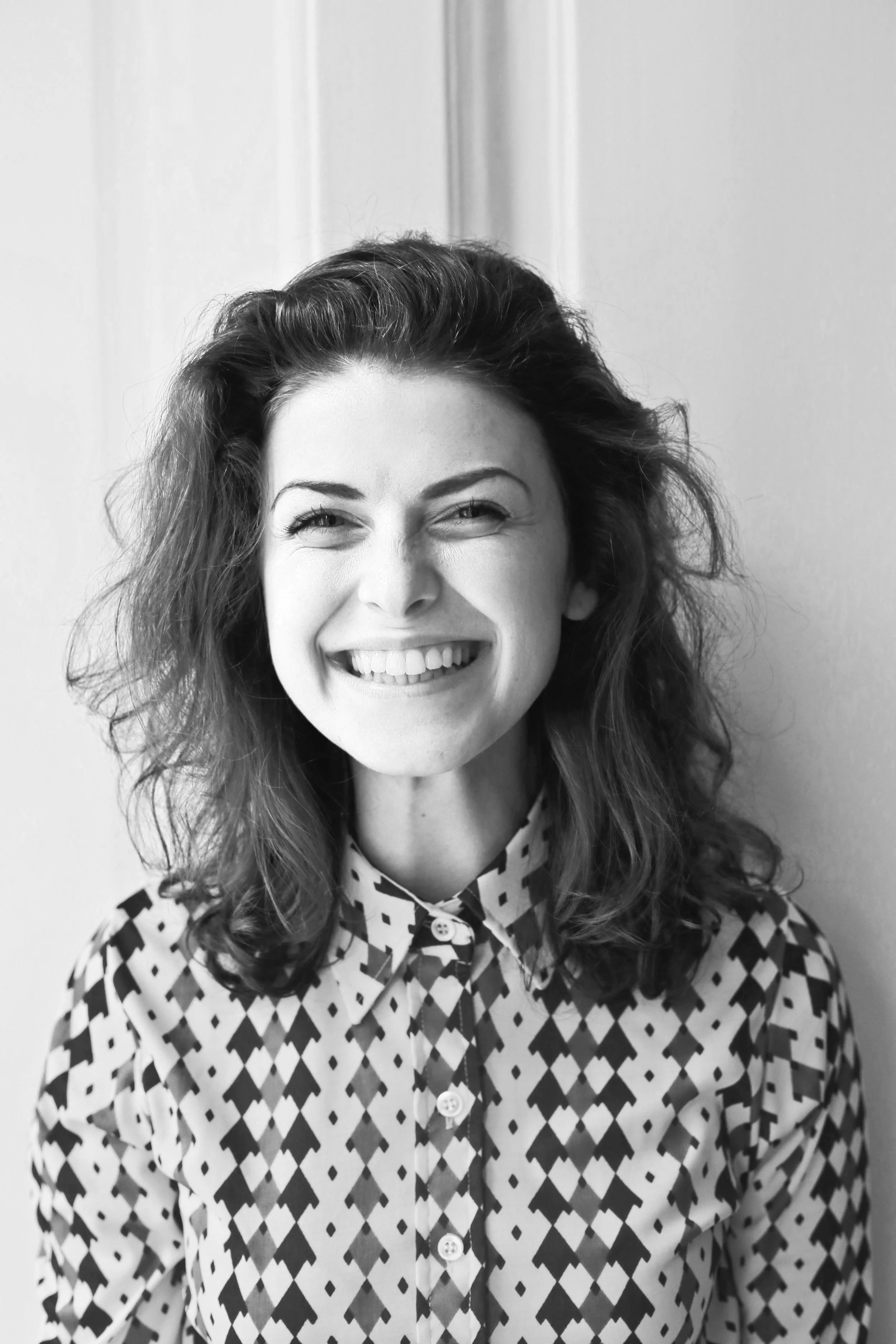 Black and white photo of a woman smiling with curly hair, wearing a patterned button-up shirt, standing against a plain wall.