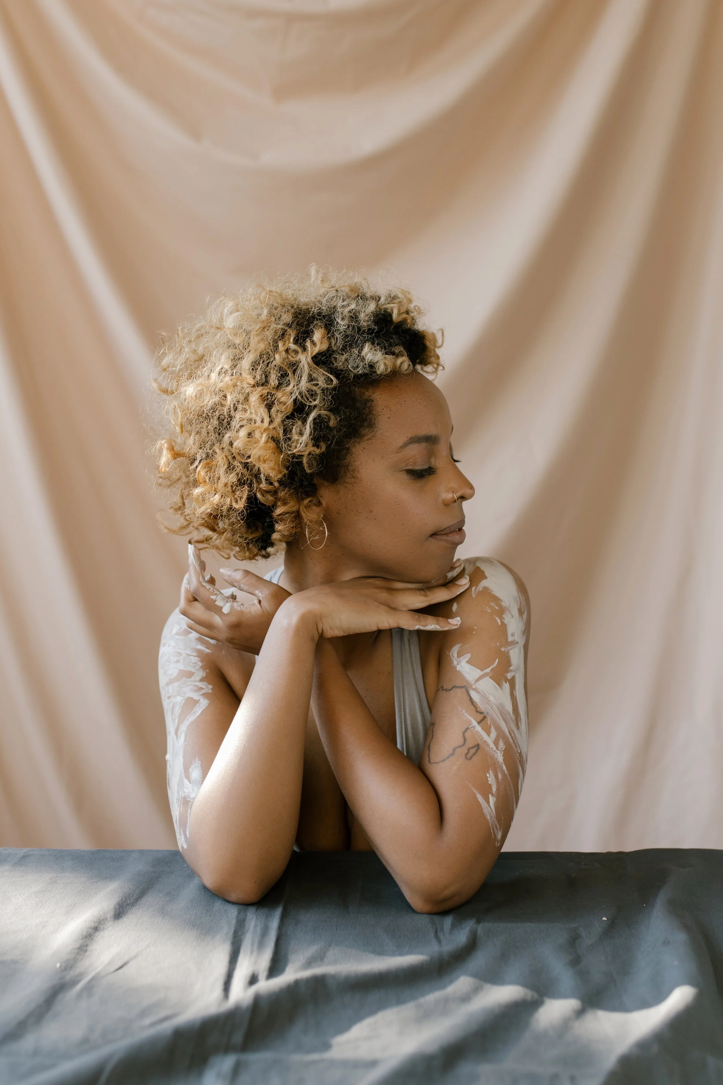 A woman with curly blonde hair, eyes closed, resting her chin on her hands, which are propped on her crossed arms on a dark surface, with a beige draped fabric background.