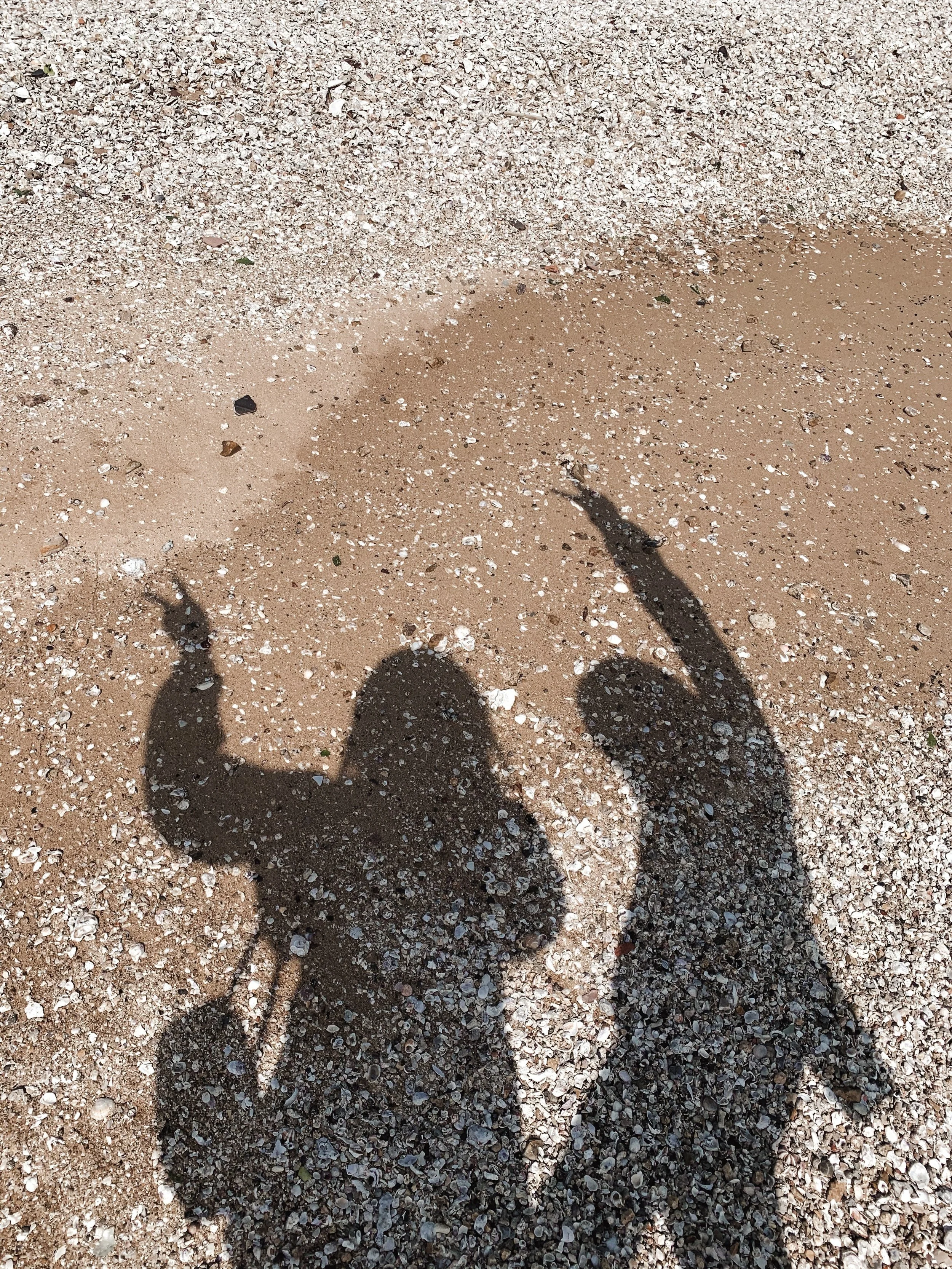 Shadows of two people making peace signs on a sandy beach with small shells and pebbles.