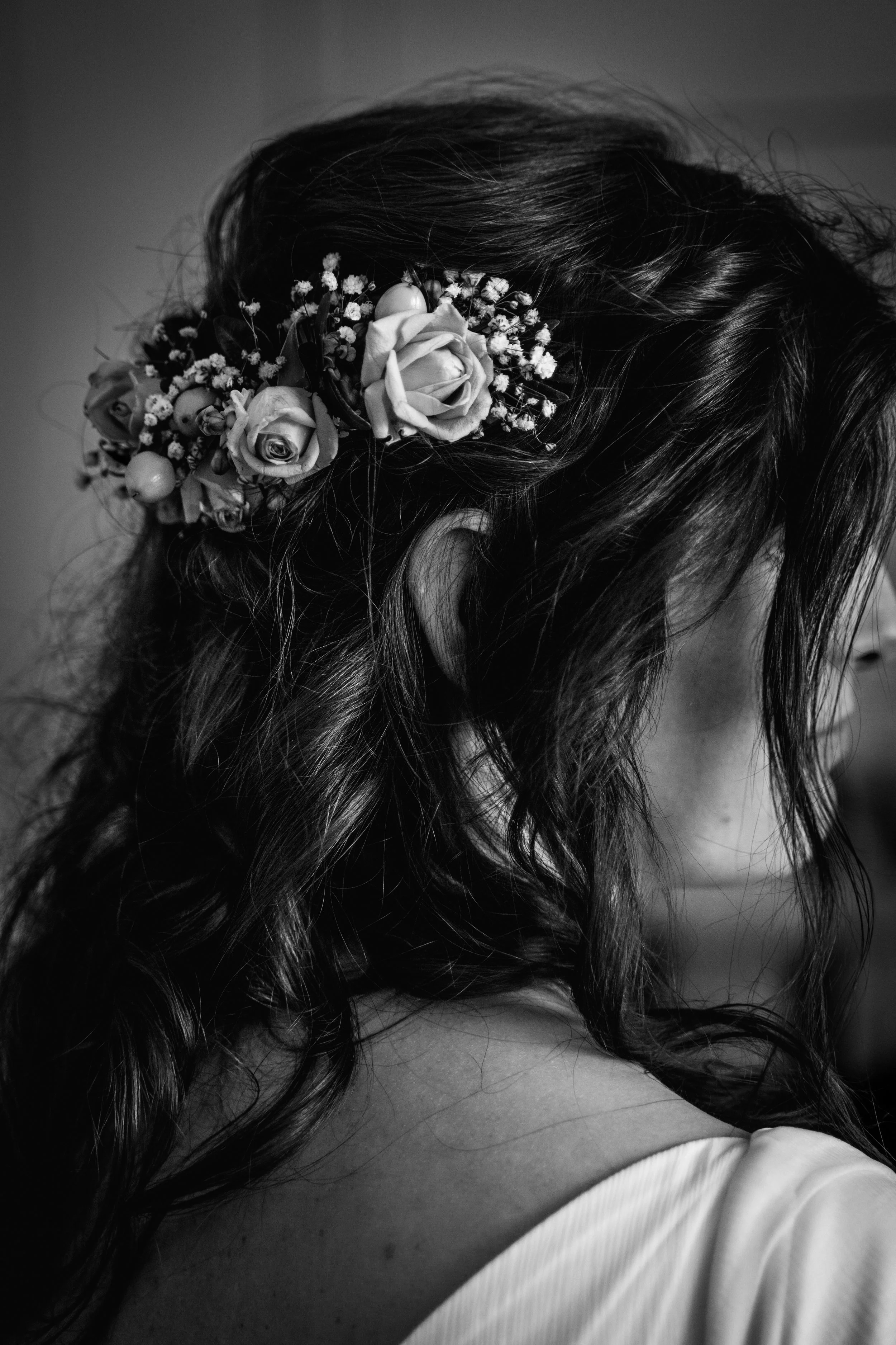 Schwarzes und weißes Nahaufnahmebild eines Mädchens mit dunklem, lockigem Haar, das eine Haarblume trägt, bestehend aus Rosen, kleinen Blumen und dekorativen Elementen.