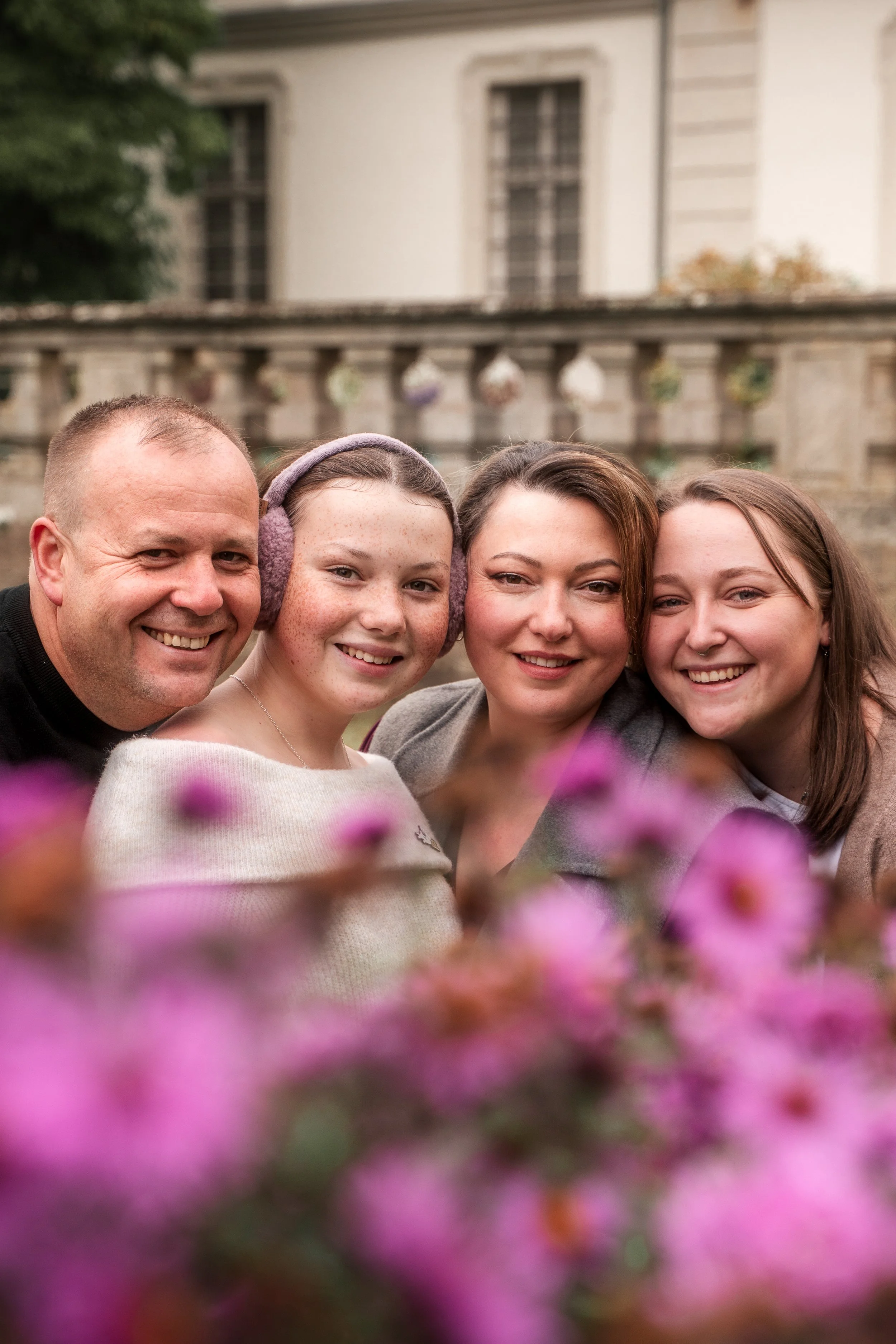 Vier Menschen lächeln in einer Nahaufnahme, im Hintergrund ein altes Gebäude. Vorn sind unscharfe lila Blumen sichtbar.