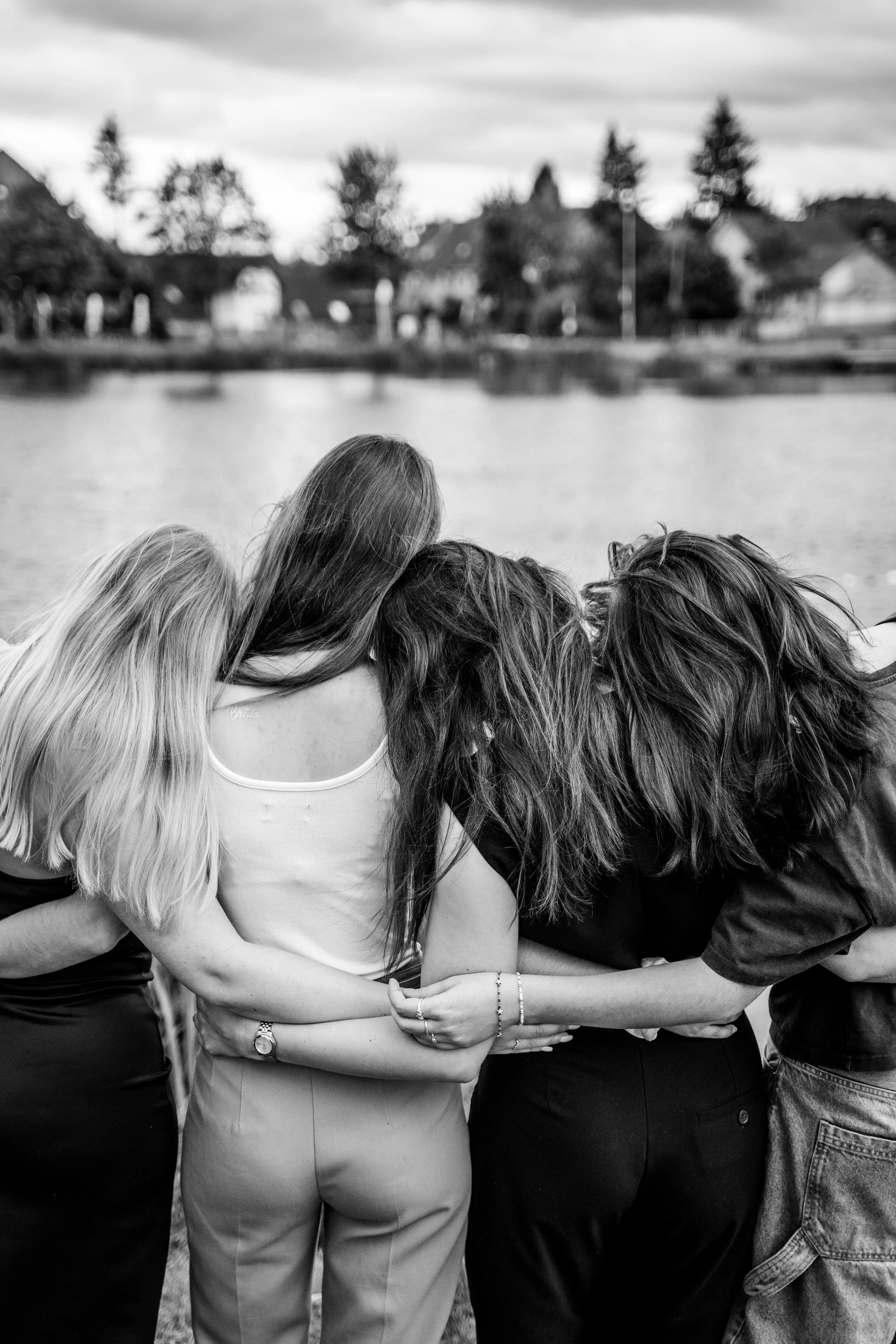Vier Frauen stehen eng beieinander am Fluss, die Köpfe aneinandergestellt, im grauen Hintergrund Häuser und Bäume, Schwarz-Weiß-Foto