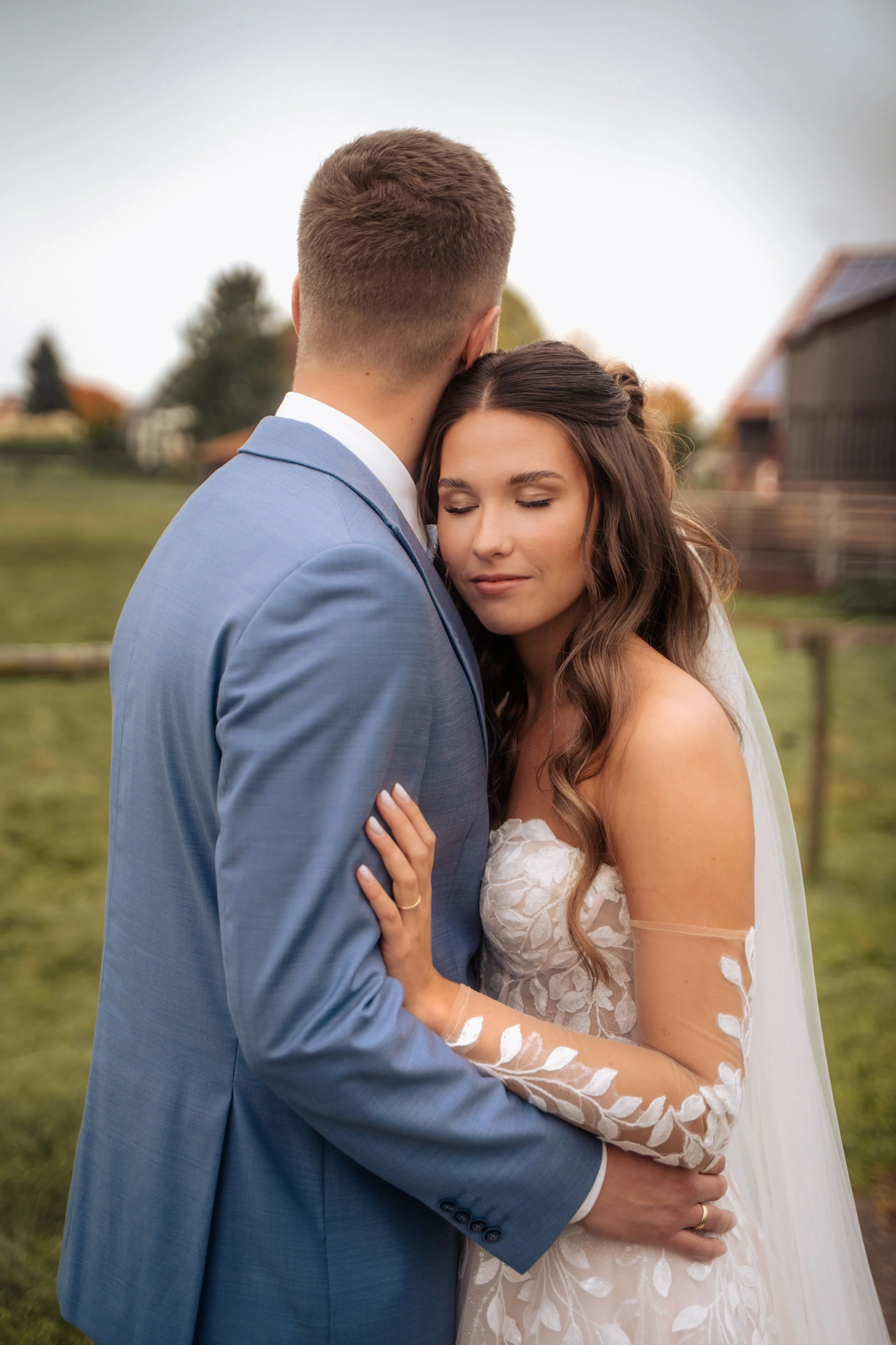 Ein Brautpaar umarmt sich im Freien bei leicht bewölktem Himmel. Der Mann trägt einen blauen Anzug und die Frau ein weißes Brautkleid mit Spitzenärmeln. Im Hintergrund sind Bäume und eine Scheune zu sehen.