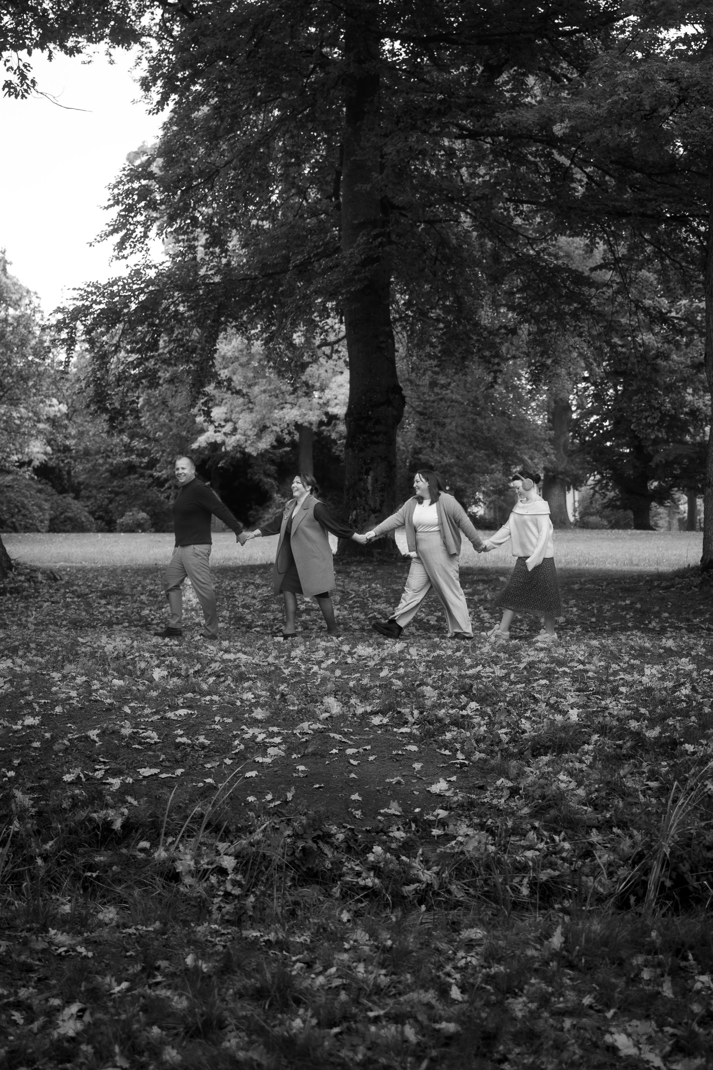 Vier Menschen laufen im Park Hand in Hand auf einem Weg, umgeben von Bäumen und im Herbst gefallene Blätter, in schwarz-weiß.