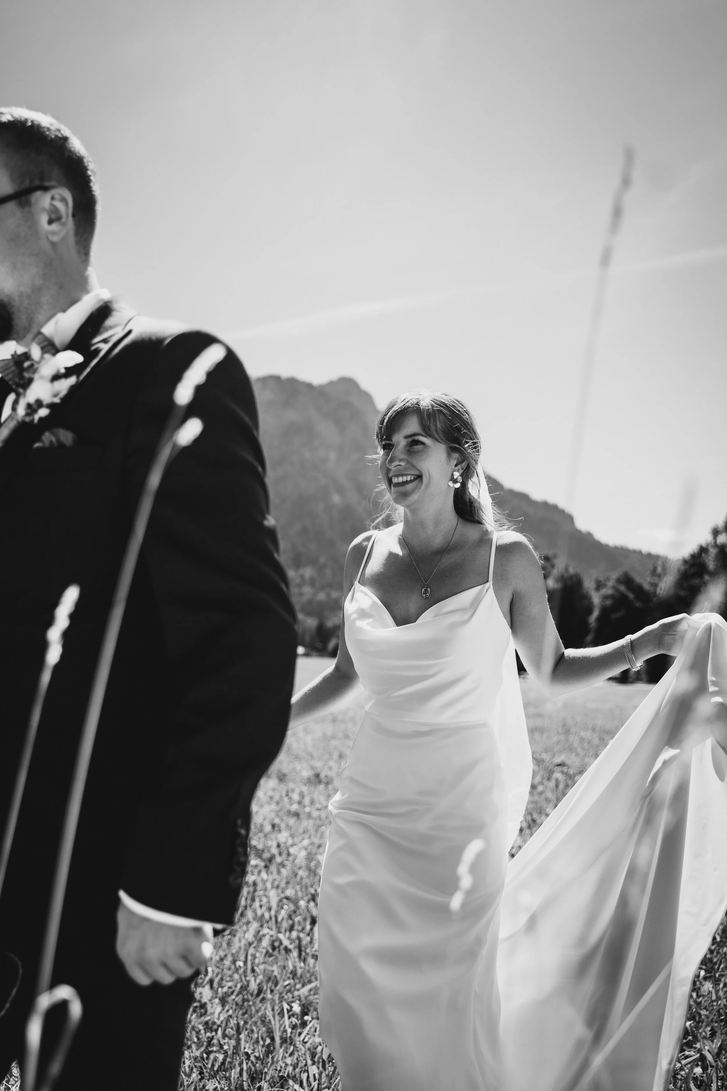 Eine lachende Frau in einem weißen Kleid bei einer Hochzeit im Freien, im Hintergrund grüne Berge, im Vordergrund eine Hand, die einen Mann in einem Anzug hält.