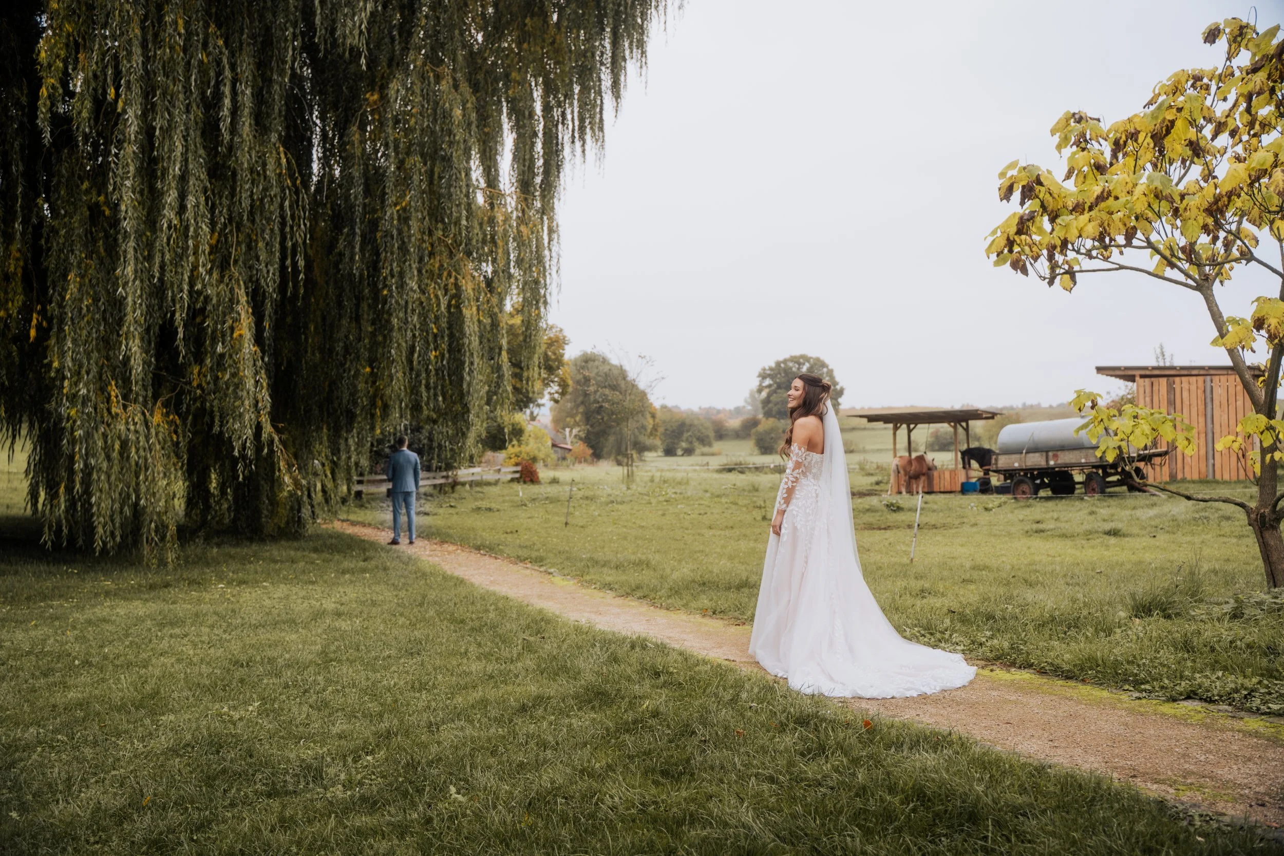 Eine Frau im weißen Hochzeitkleid steht auf einem ländlichen Feld bei einem Baum, während ein Mann im Anzug entfernt auf einem Weg geht. Es gibt landwirtschaftliche Geräte und ein kleines Holzgebäude im Hintergrund.