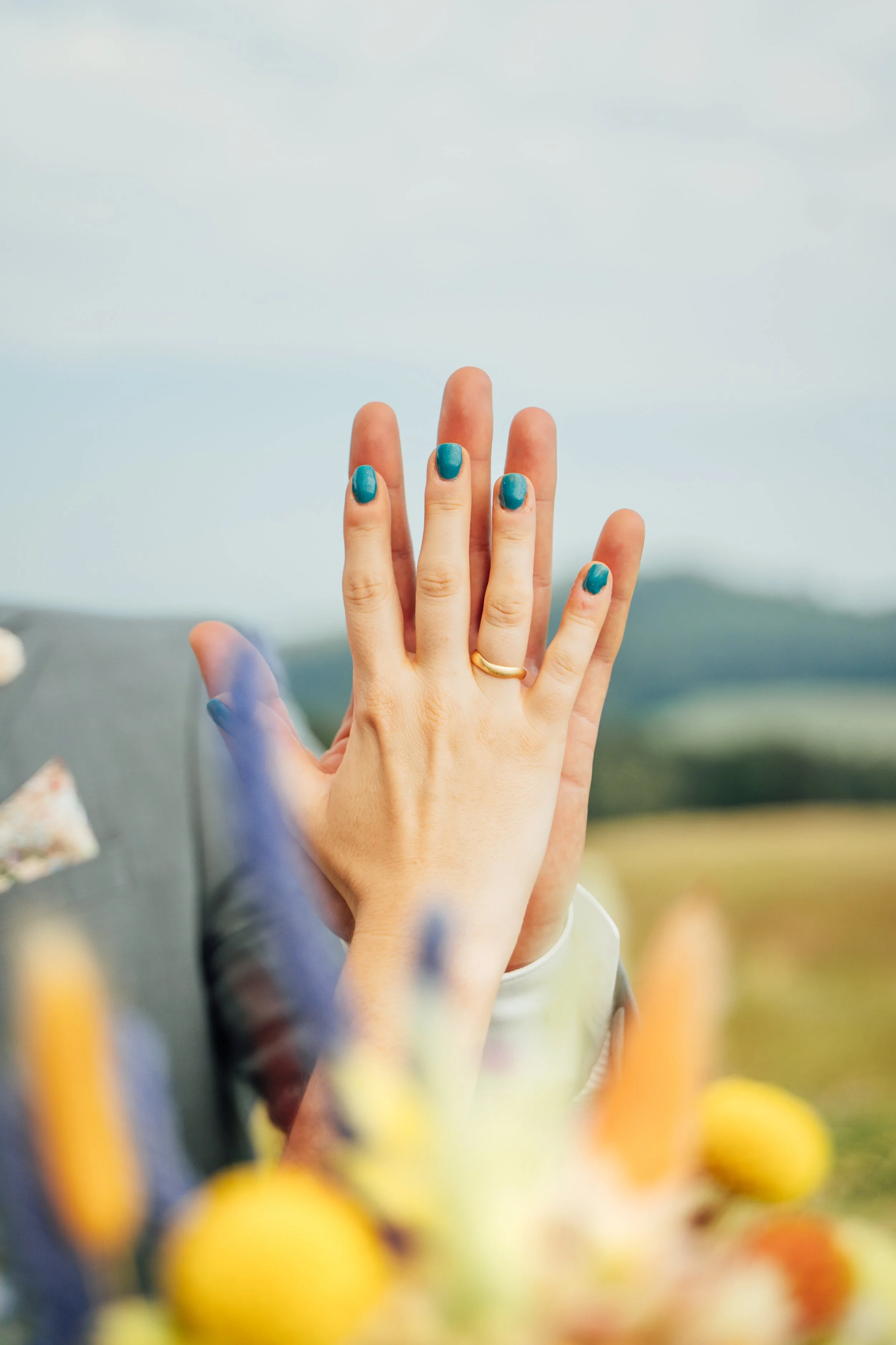 Hände eines Menschen mit aufgemalten blauen Fingernägel, die sich berühren, im Hintergrund eine Landschaft mit Himmel. Im Vordergrund sind ausgeblichene gelbe und orange Früchte oder Blumen.