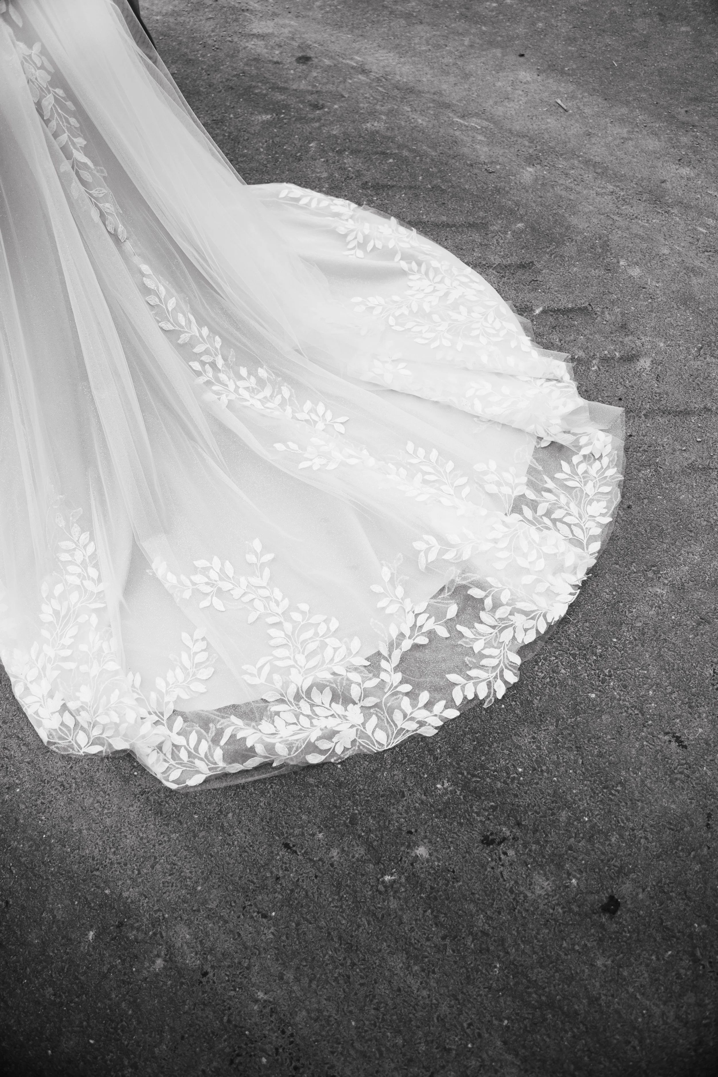 Ein Teil eines weißen, eleganten Brautkleids mit zarten Blätterstickereien, liegt auf einer dunklen Asphaltfläche.