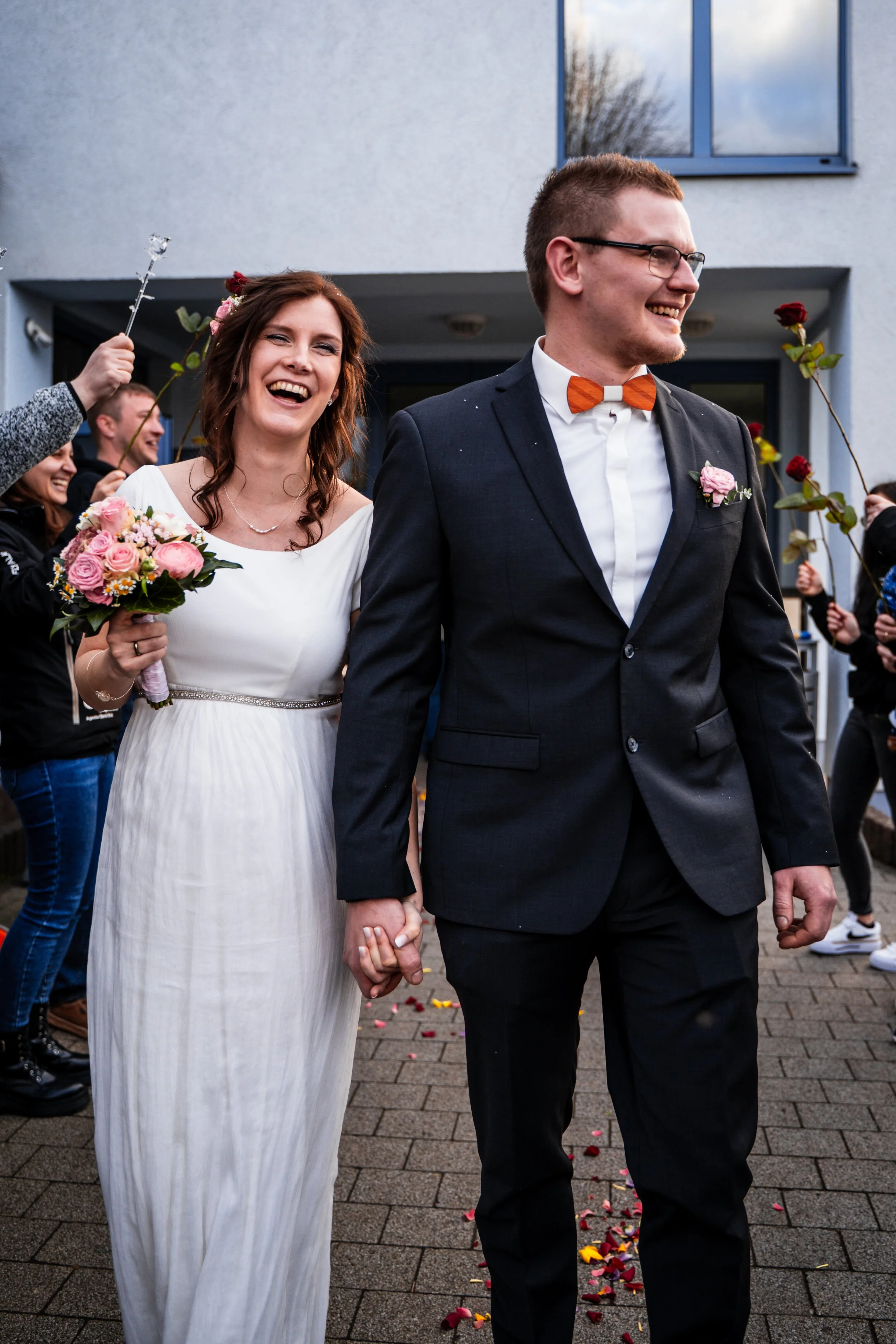 Ein lachendes Brautpaar hält Händchen, während sie an einer Hochzeit teilnehmen, umgeben von Freunden, die Rosen und Blütenblätter werfen.