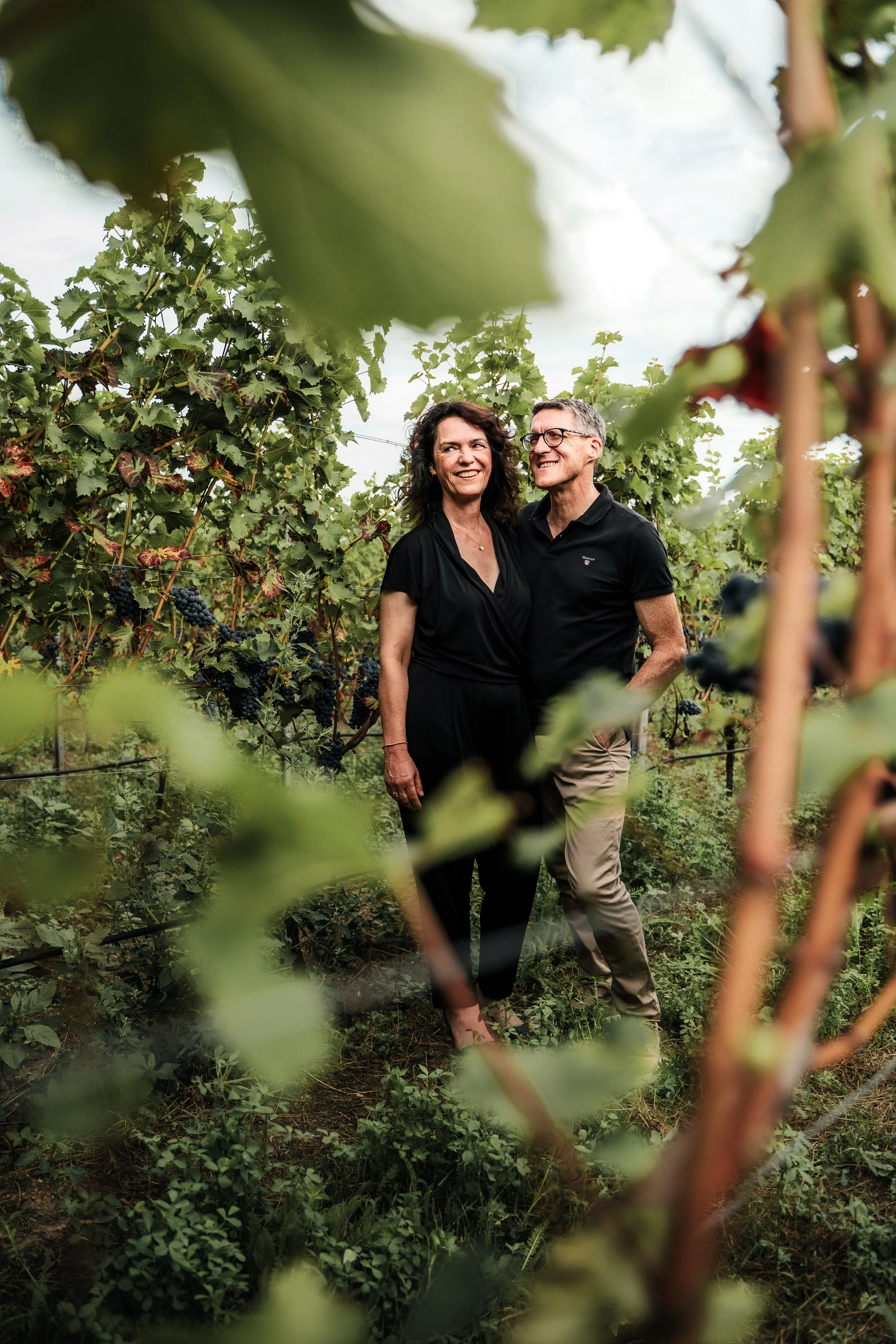 Ein Mann und eine Frau stehen in einem Weinberg, umgeben von Reben und Trauben, während sie miteinander lachen.