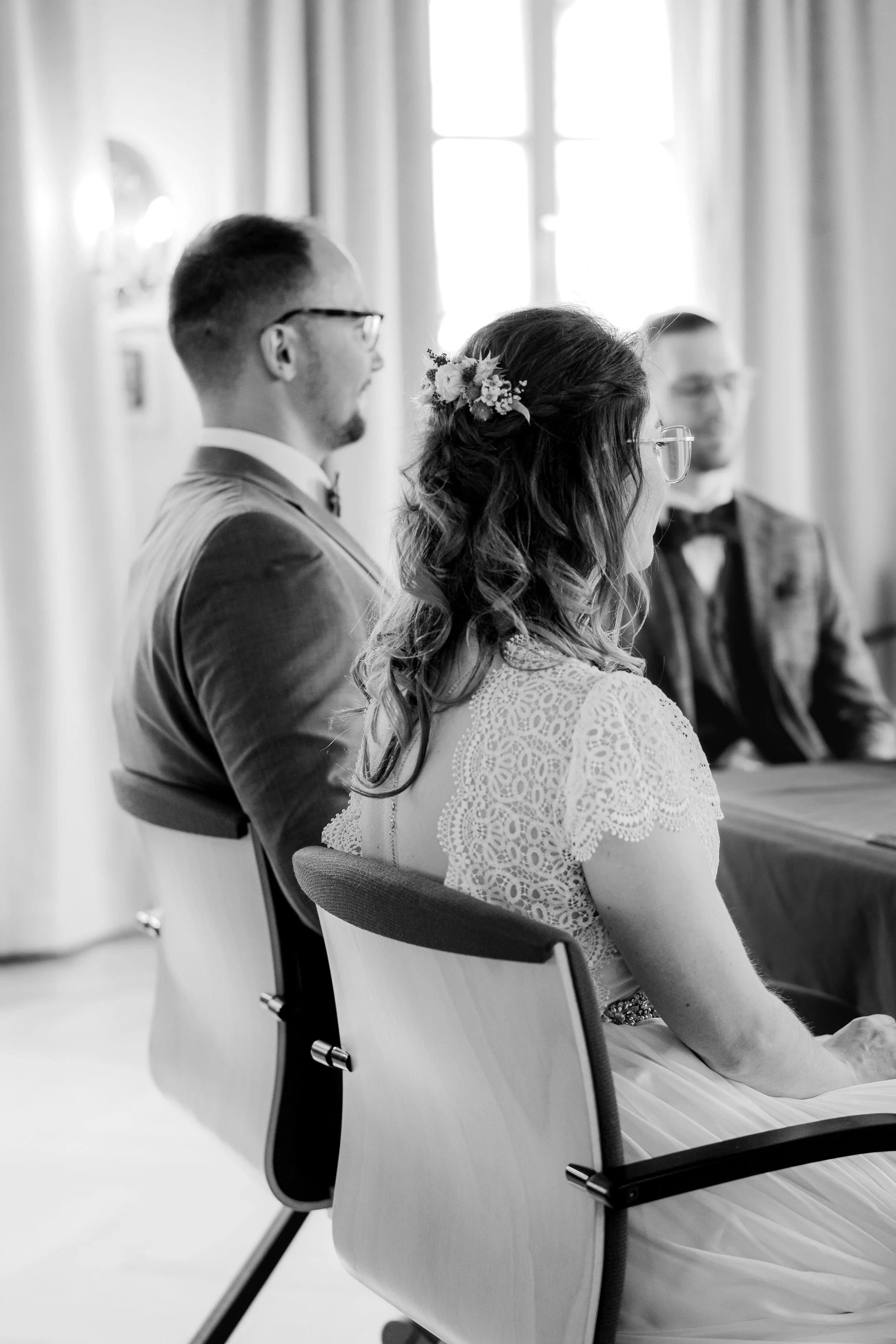 Schwarz-weiß-Foto eines Brautpaares bei einer Zeremonie, sitzend auf Stühlen in einem hell erleuchteten Raum, die Braut trägt eine weiße Spitze Brautkleid mit Haarschmuck, die beiden Männer tragen Anzüge mit Fliege, im Hintergrund Gardinen vor einem 