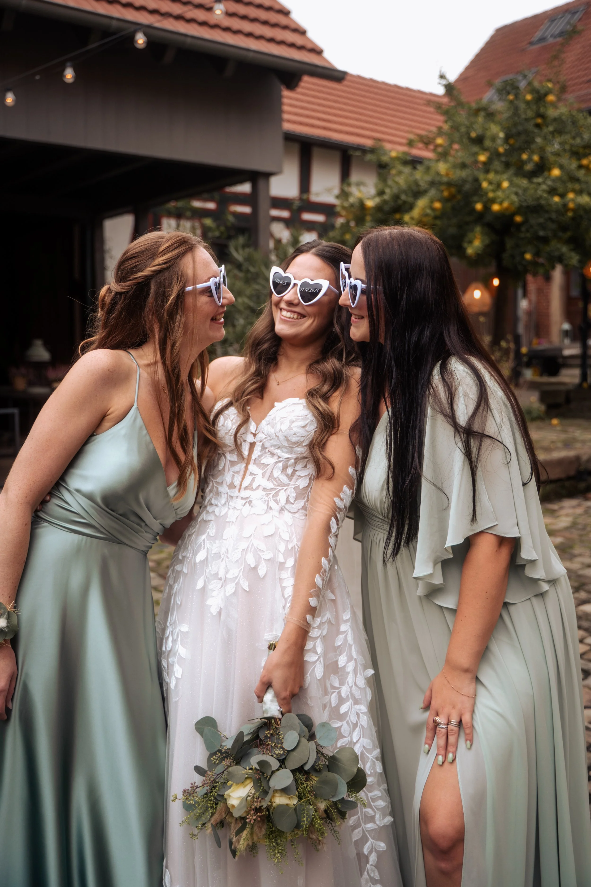 Drei Frauen bei einer Hochzeit, alle tragen Sonnenbrillen mit Herzdesign, die Mittefrau trägt ein weißes Brautkleid, die anderen zwei Frauen tragen grüne Kleider, im Hintergrund Häuser und einen Apfelbaum.