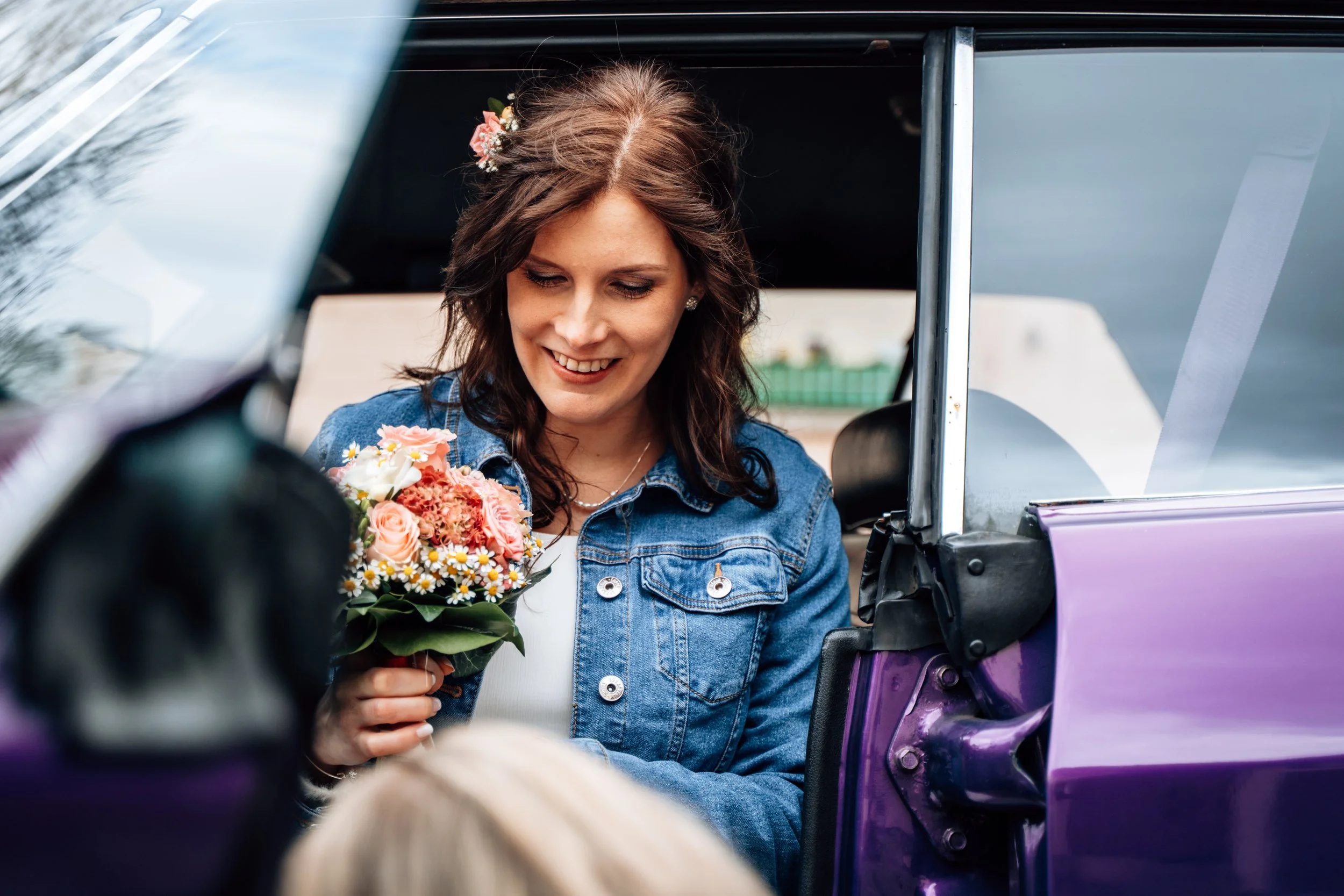 Eine junge Frau mit braunen, gewellten Haaren und Blumen im Haar, lächelnd, hält einen Blumenstrauß in der Hand, während sie in einem älteren, offenen Auto sitzt, möglicherweise auf dem Weg zu einer Feier.
