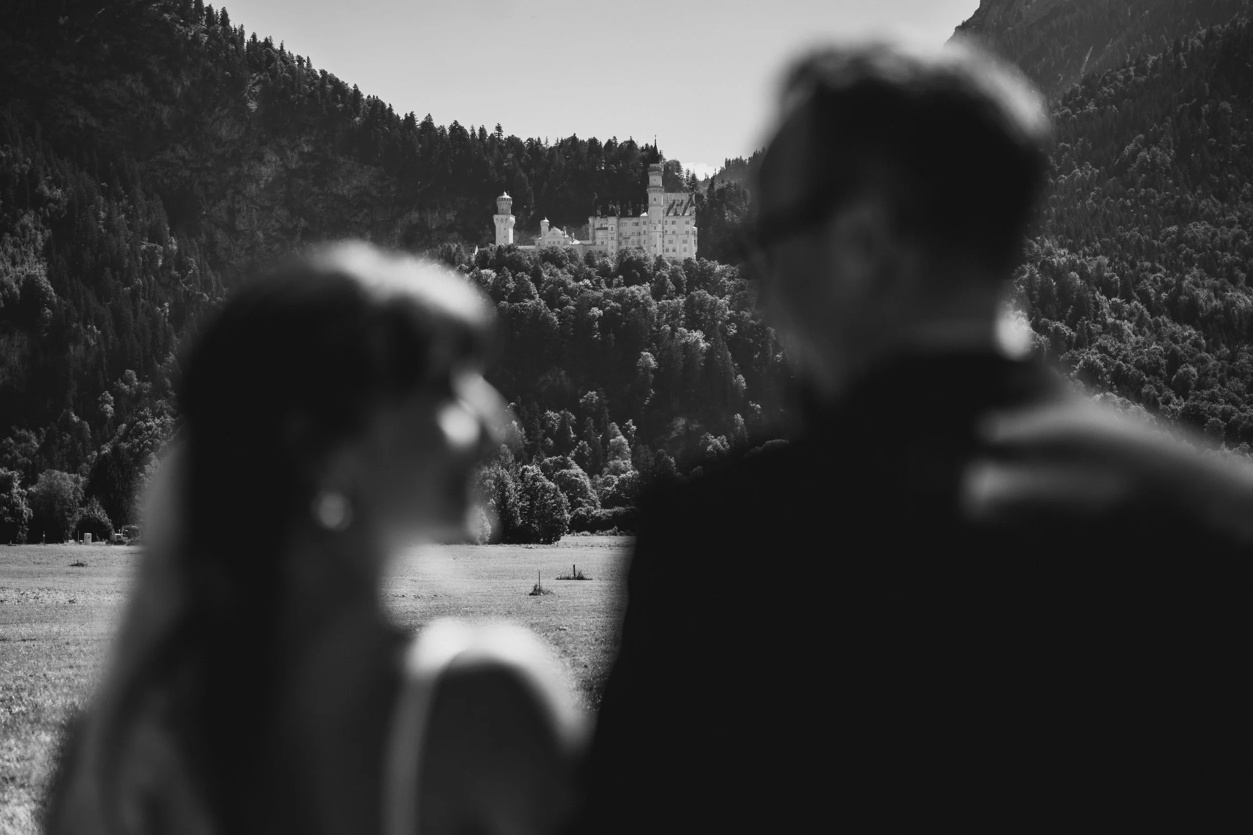 Schwarz-weiße Aufnahme eines Schlosses auf einem Hügel, im Vordergrund sind die Köpfe von mehreren Menschen, die das Schloss im Hintergrund betrachten.