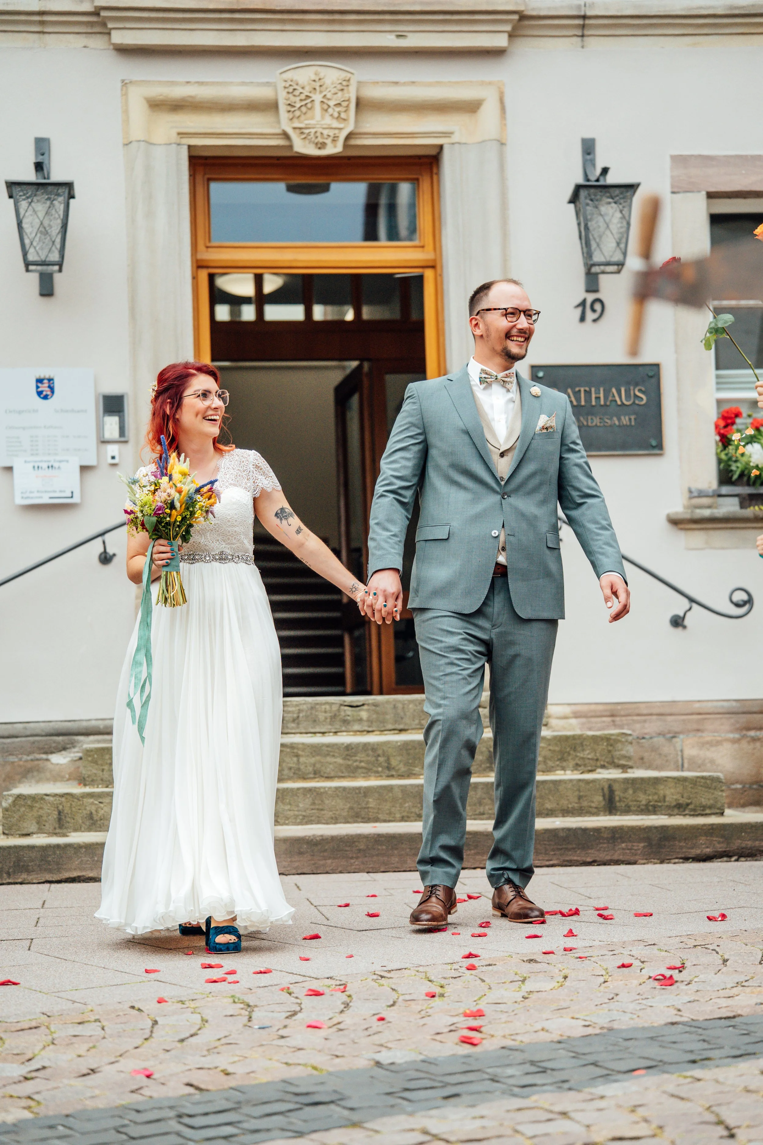 Ein glückliches Paar hält sich an den Händen, vor einer Kirche, nach einer Heirat, auf Steinstufen, mit roten Rosenblättern auf dem Boden. Der Mann trägt einen grauen Anzug, Brille und Fliege, die Frau trägt ein weißes Hochzeitskleid und hat eine Blu