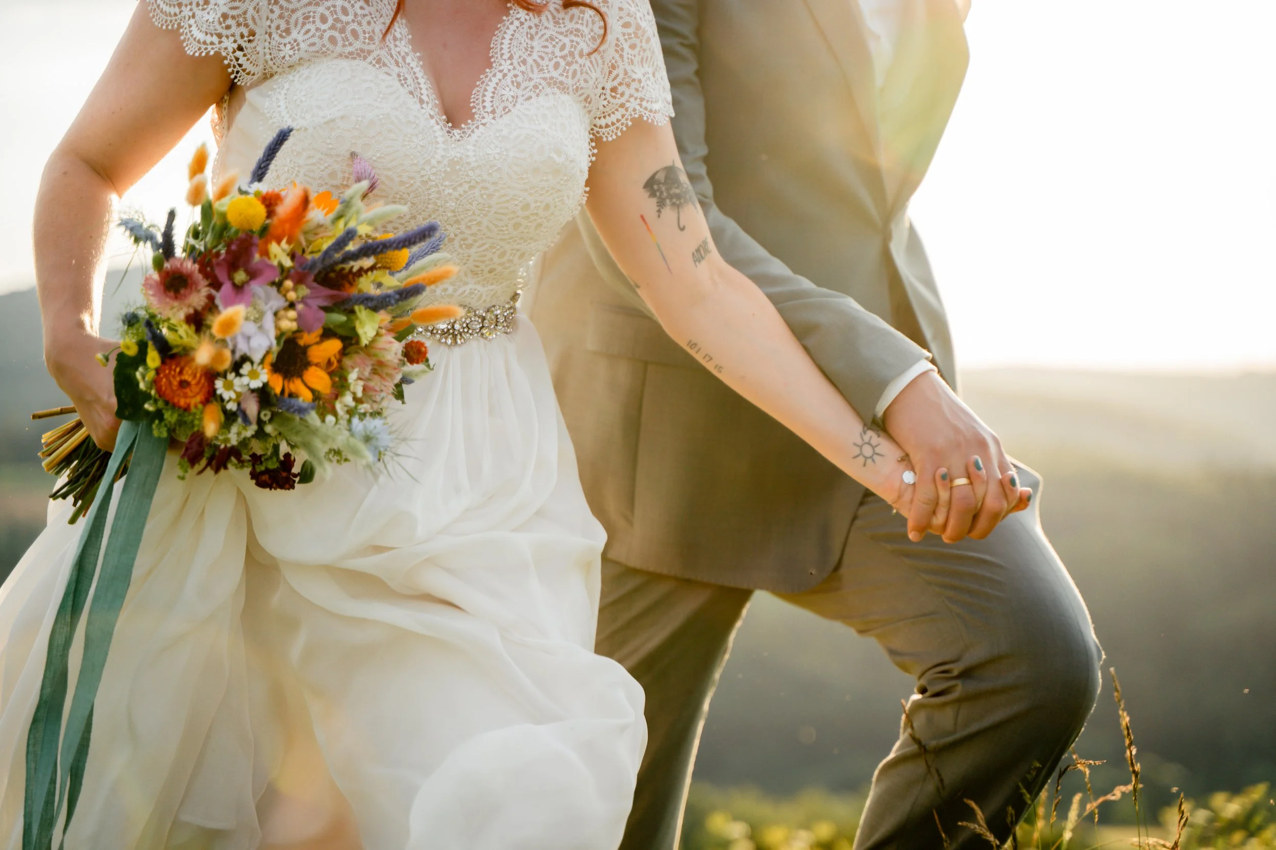 Nahaufnahme eines Paares bei einer Hochzeit, das Händchen hält. Die Braut trägt ein Spitzenkleid mit Spaghettiträgern und einen Blumenstrauß, während der Bräutigam einen grauen Anzug trägt.