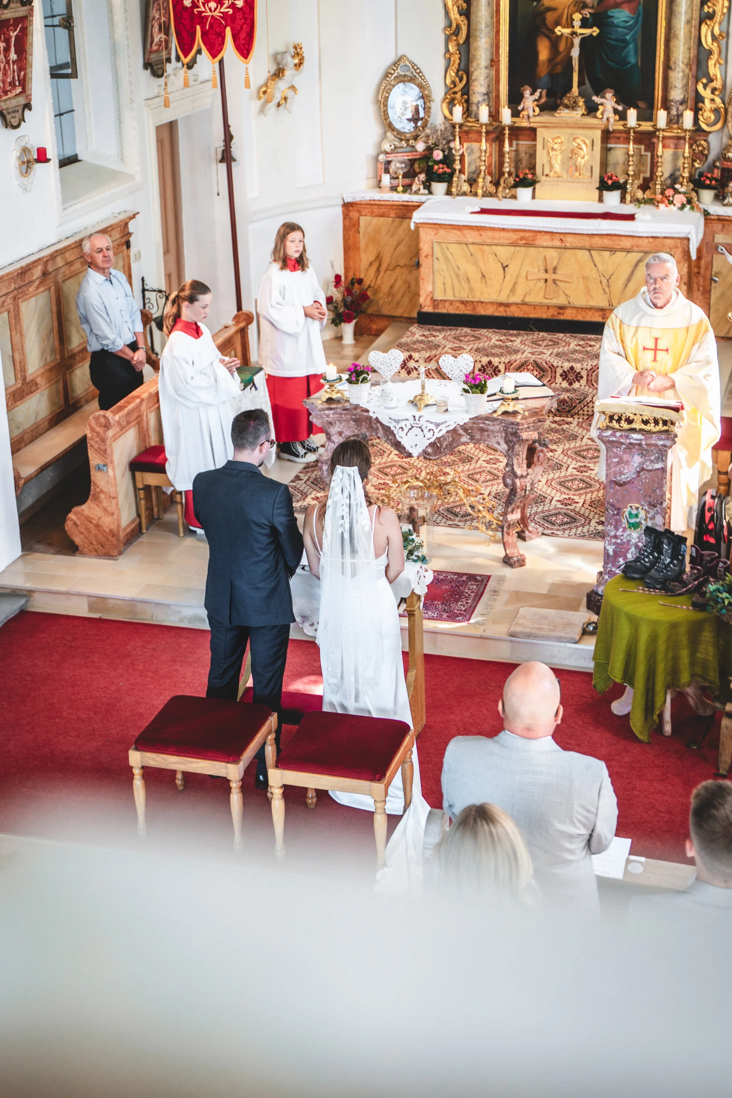 Ein Hochzeitspaar steht vor dem Altar in einer Kirche während einer Zeremonie, mit Angehörigen und Gästen im Hintergrund.