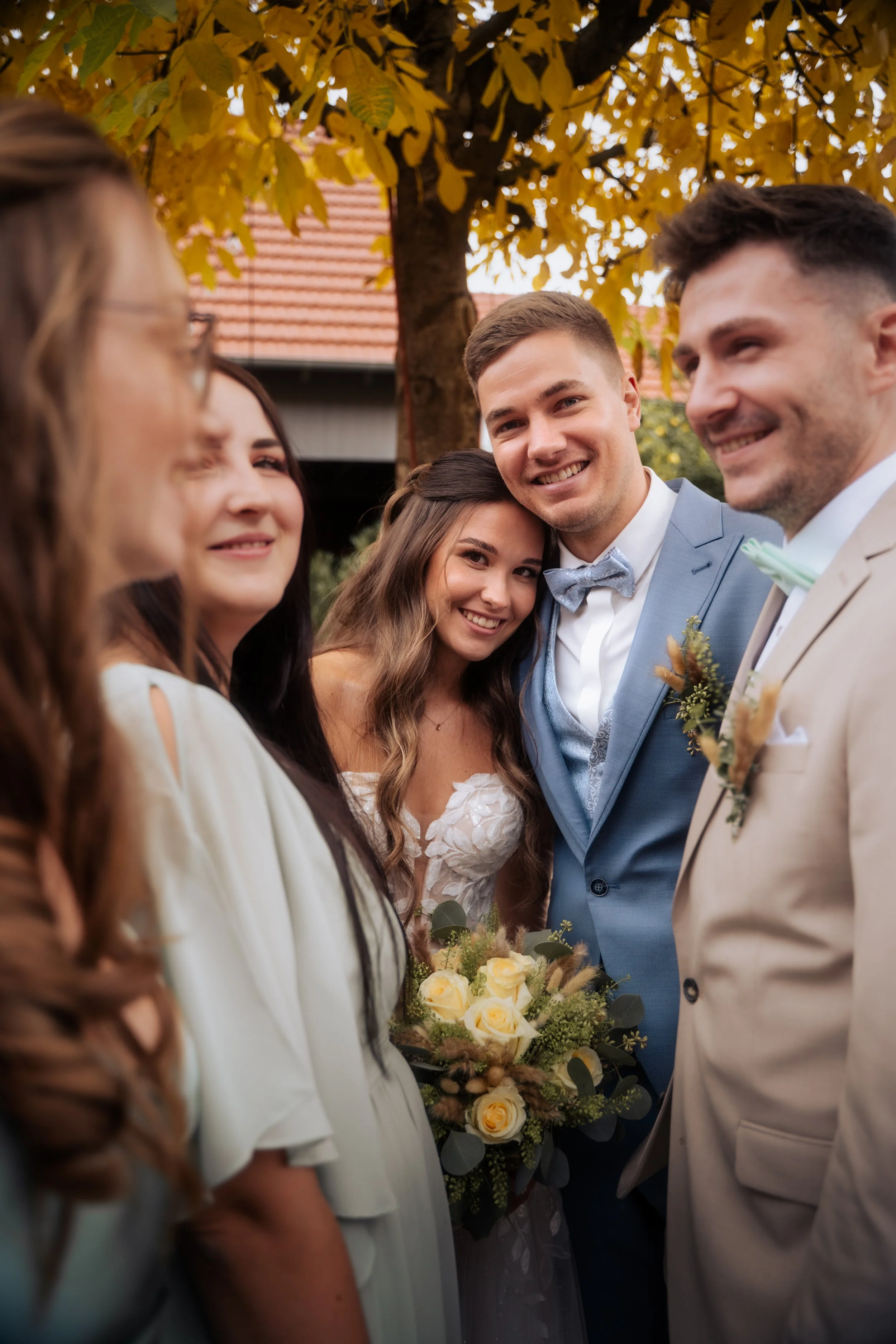 Eine Gruppe von Menschen bei einer Hochzeitsfeier im Freien, umgeben von Bäumen mit herbstlichen Blättern. Einige tragen helle Kleider und haben ihre Arme um die Braut und den Bräutigam, die lächelnd in den Mittelpunkt gestellt sind.