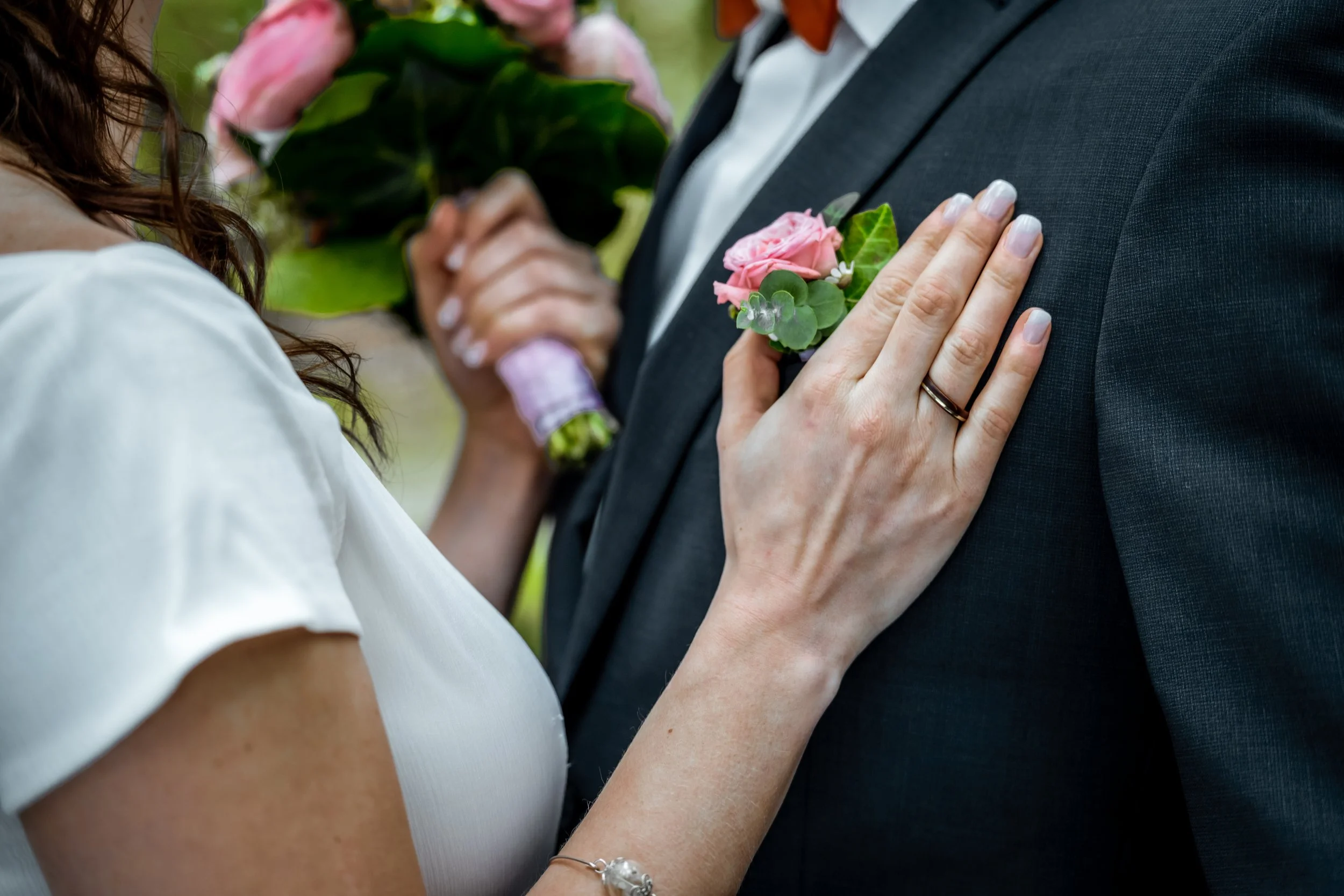 Ein Paar hält sich bei einer Hochzeit an den Händen, während die Braut ihre Hand auf die Brust des Bräutigams legt. Beide tragen formelle Kleidung und die Frau trägt ein Ring am Finger. Es gibt Blumen im Hintergrund.