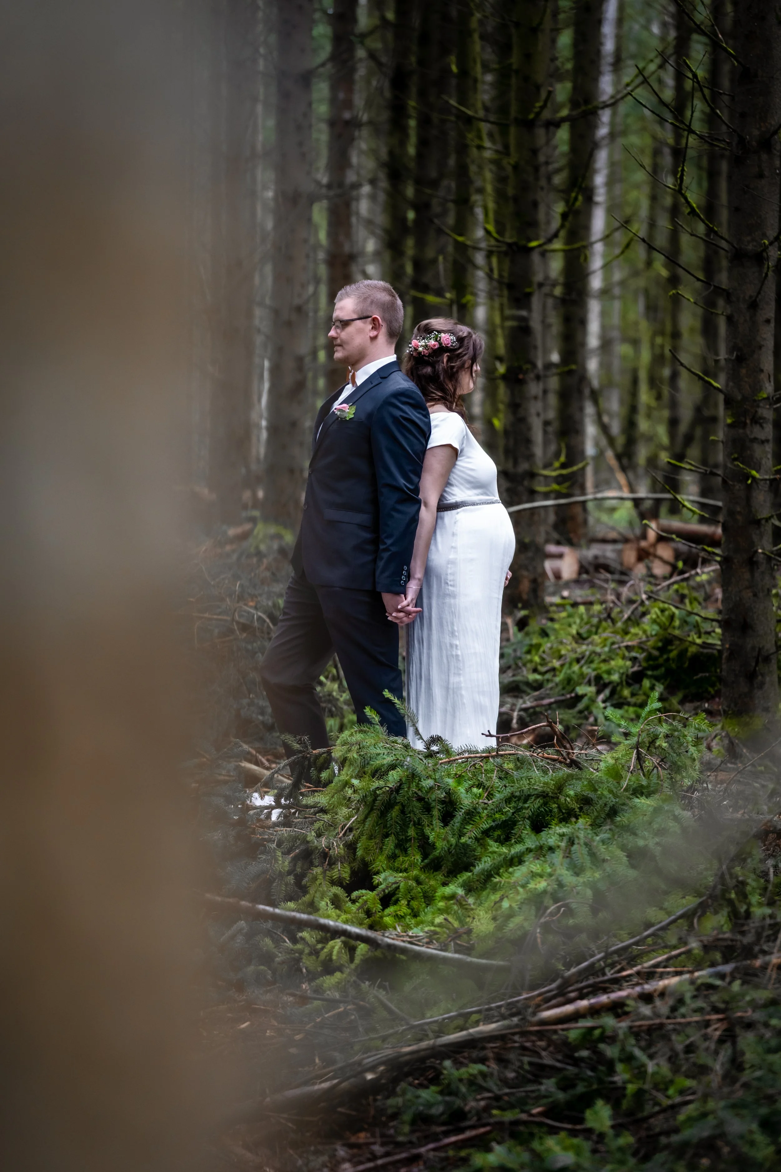 Ein Paar, ein Mann und eine Frau, stehen in einem Wald. Der Mann trägt einen dunklen Anzug und eine Fliege, die Frau trägt ein weißes Kleid und eine Blumenkrone im Haar. Sie halten Händchen, stehen Rücken an Rücken und blicken nach vorne.