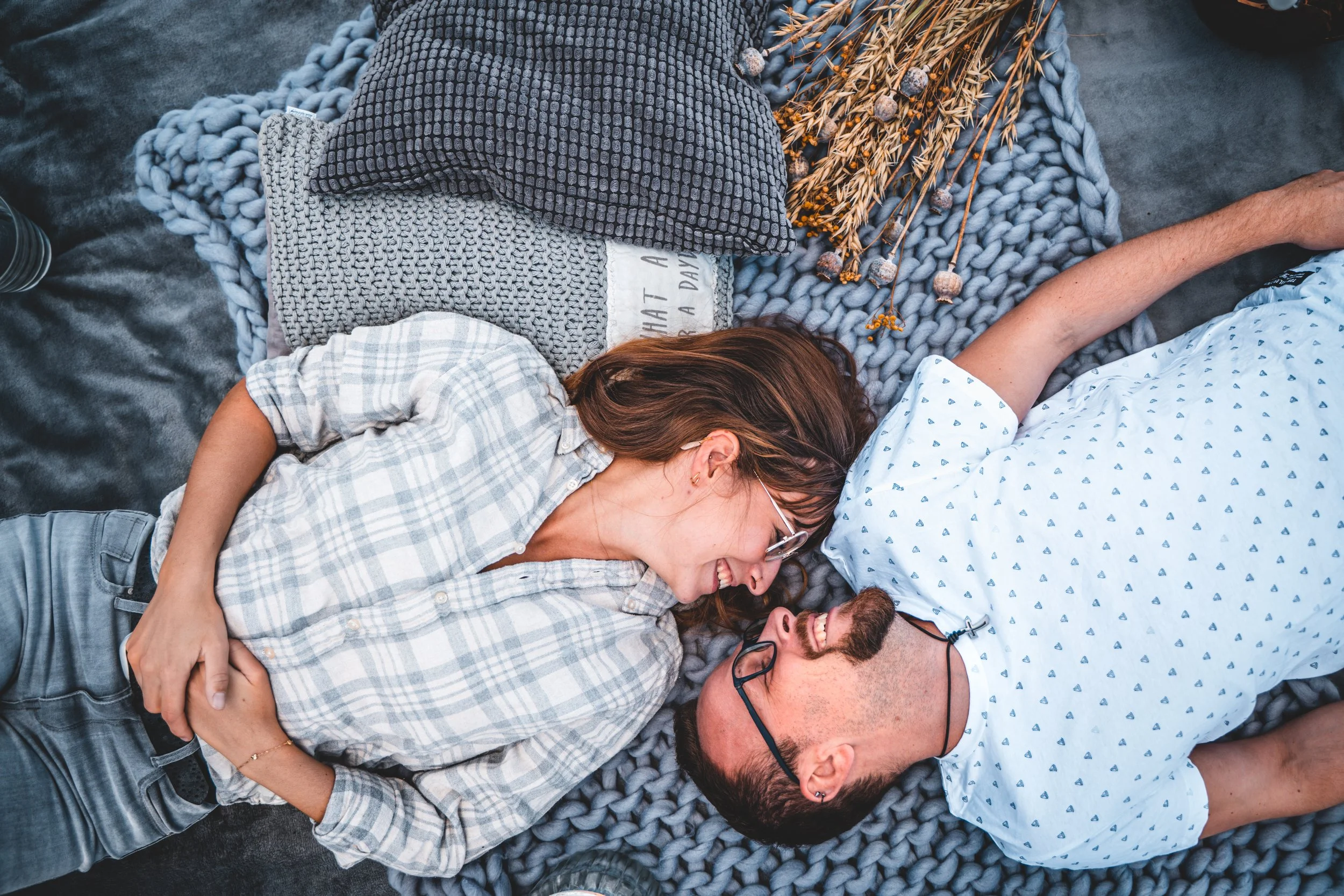 Zwei Menschen, ein Mann mit Brille und ein Frau mit Locken, liegen im Bett und lachen, während sie sich umarmen.