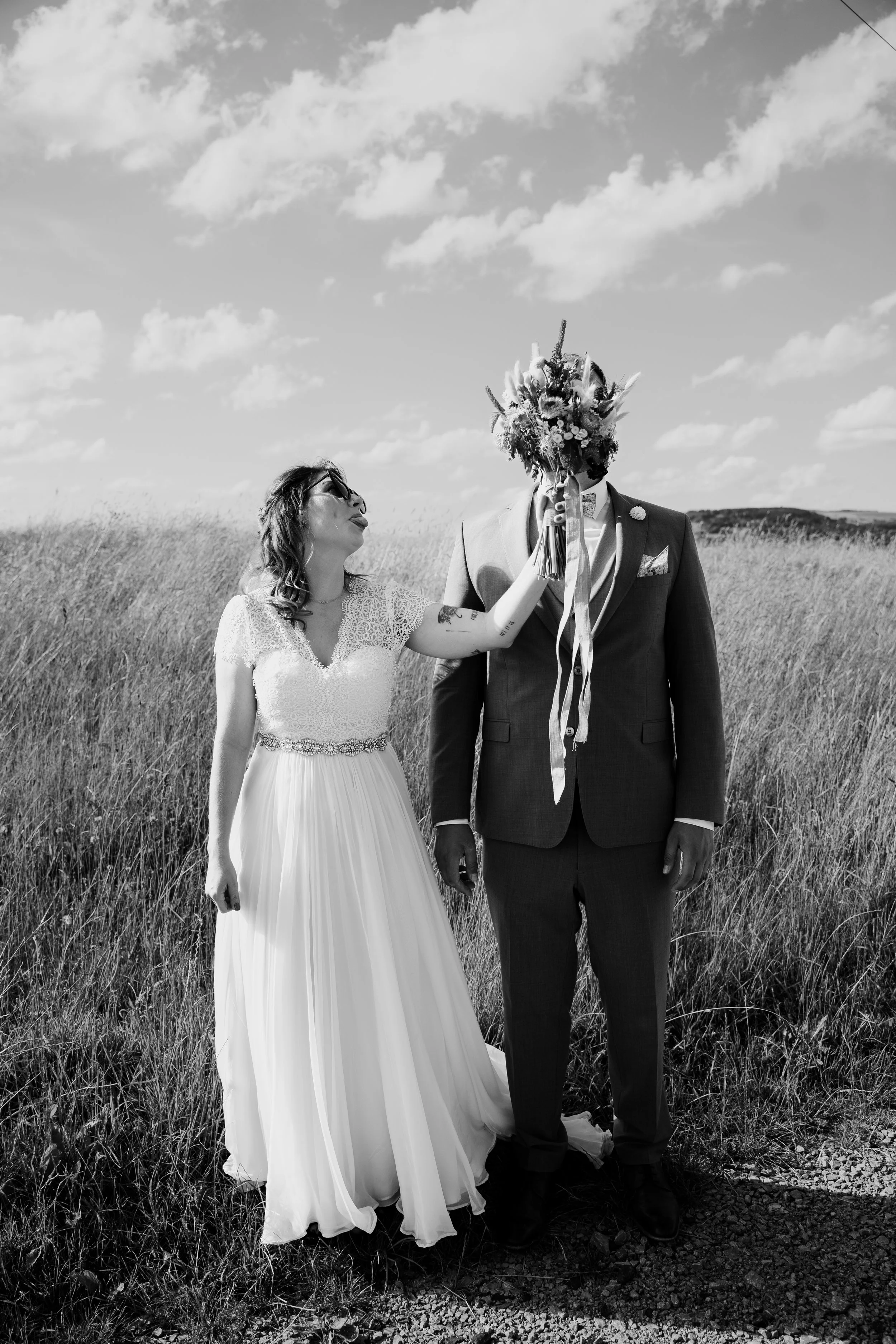 Ein Brautpaar steht in einem Feld. Die Braut trägt ein weißes Hochzeitskleid mit Spitze, der Bräutigam trägt einen dunklen Anzug. Das Brautpaar hält gemeinsam einen großen Brautstrauß, der die Sicht des Bräutigams verdeckt. Der Himmel ist mit Wolken 