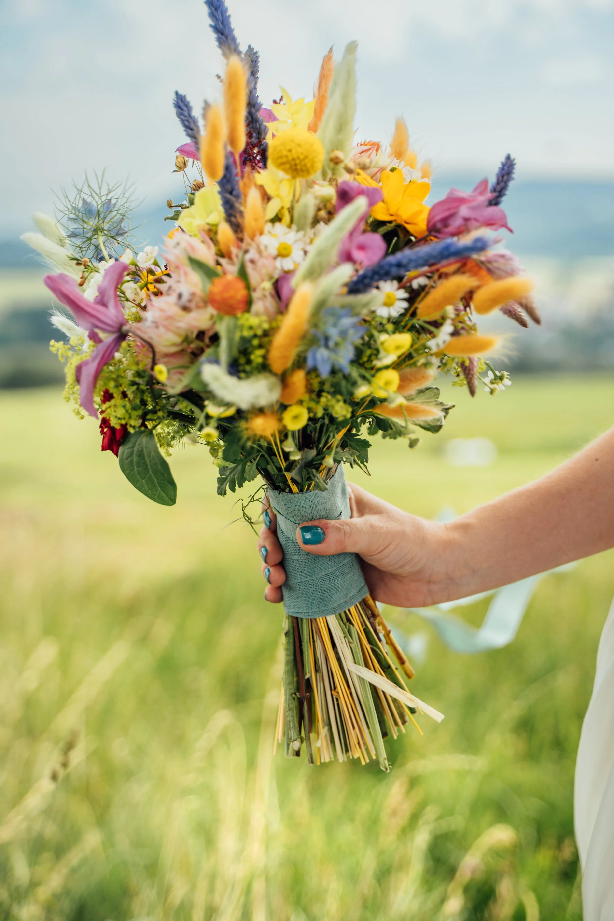 Hand hält einen bunten Blumenstrauß vor grünem, offener Feldhintergrund.