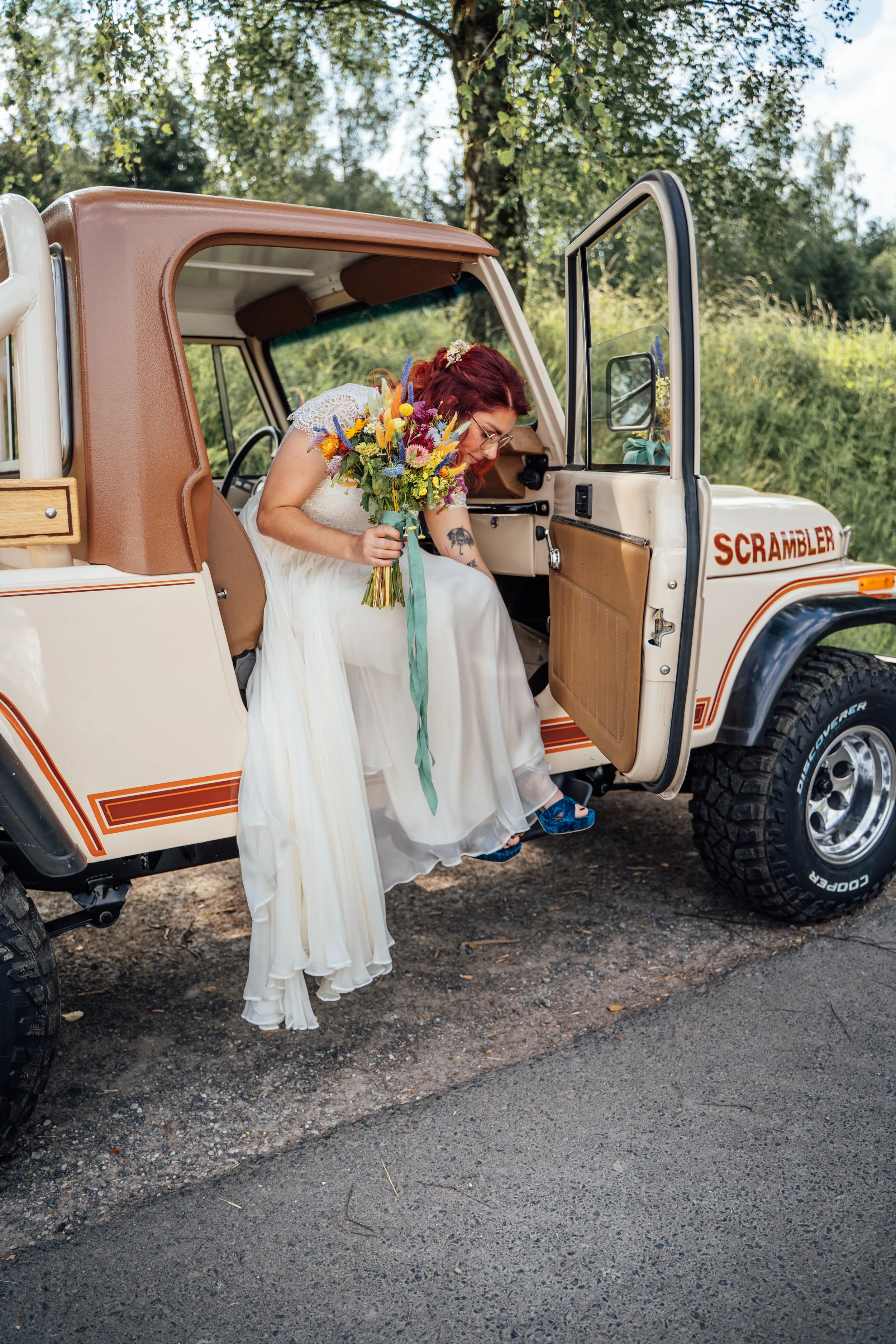 Eine Frau mit rotem Haar, Brille und Hochzeitskleid sitzt in einem Jeep, hält einen Blumenstrauß und schaut nach unten.