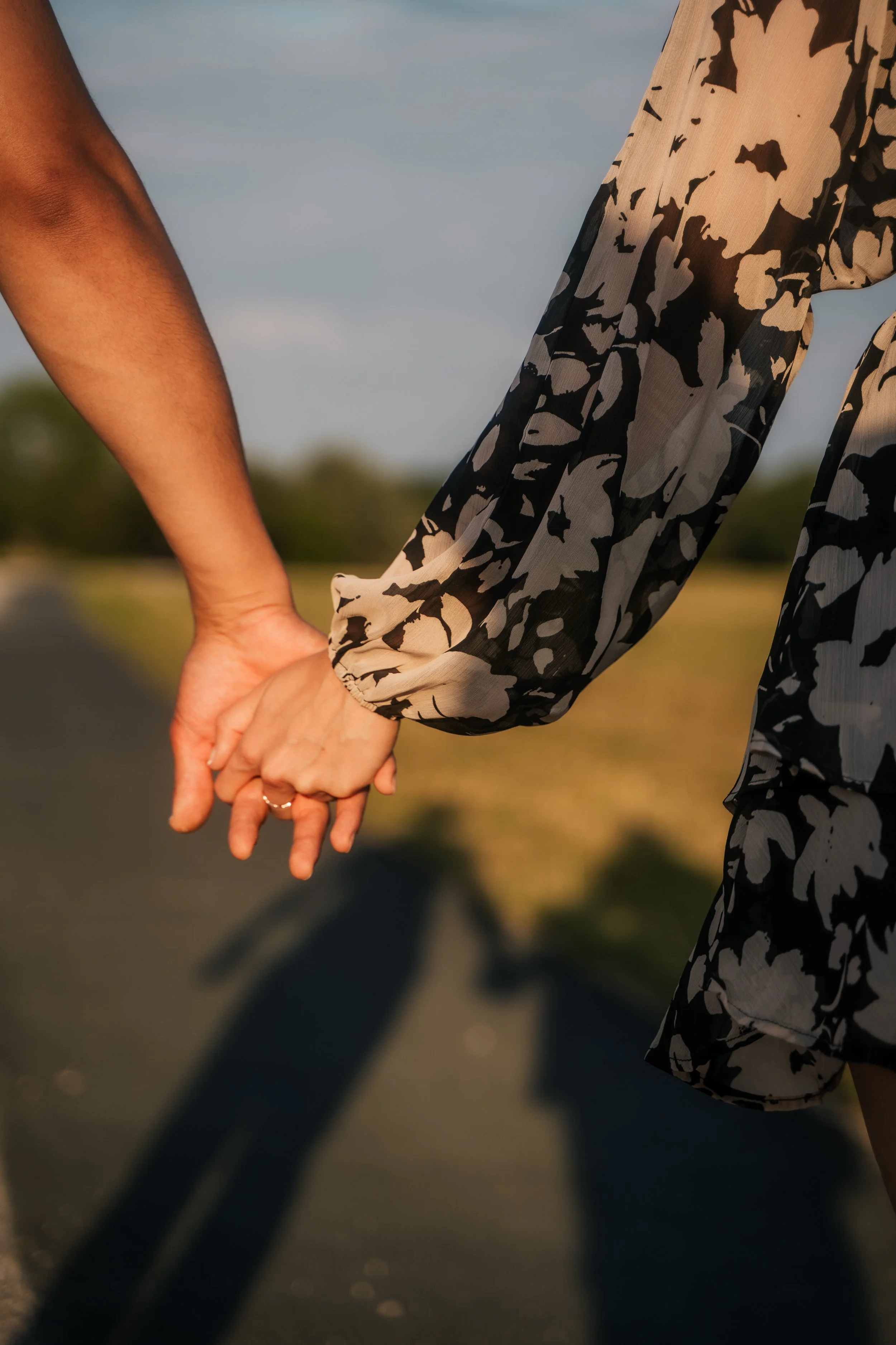 Nahaufnahme von zwei Personen, die sich an den Händen halten, bei Sonnenuntergang. Eine Person trägt ein schwarz-weiß gemustertes Kleid, die andere Person hat einen unbekannten Kleidungsstil. Der Schatten ihrer Hände wird auf die Straße geworfen.
