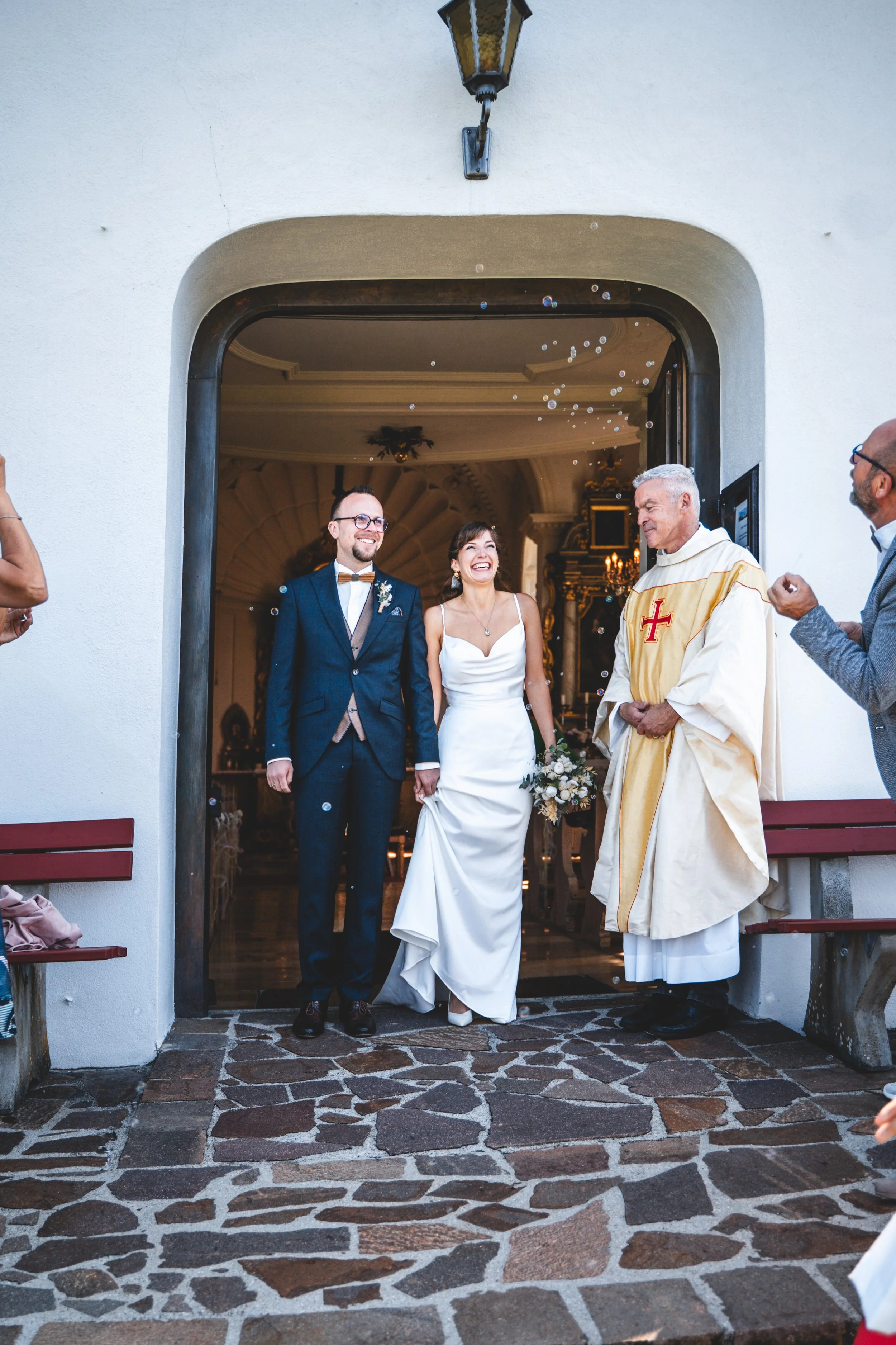 Ein frisch vermähltes Brautpaar verlässt die Kirche, umgeben von Freunden und Familie beim Hochzeitstag. Das Brautpaar lächelt, während der Pfarrer in heller Robe mit Kreuz auftritt. Draußen ist der Eingang zur Kirche sichtbar, mit Steinen auf dem Bo